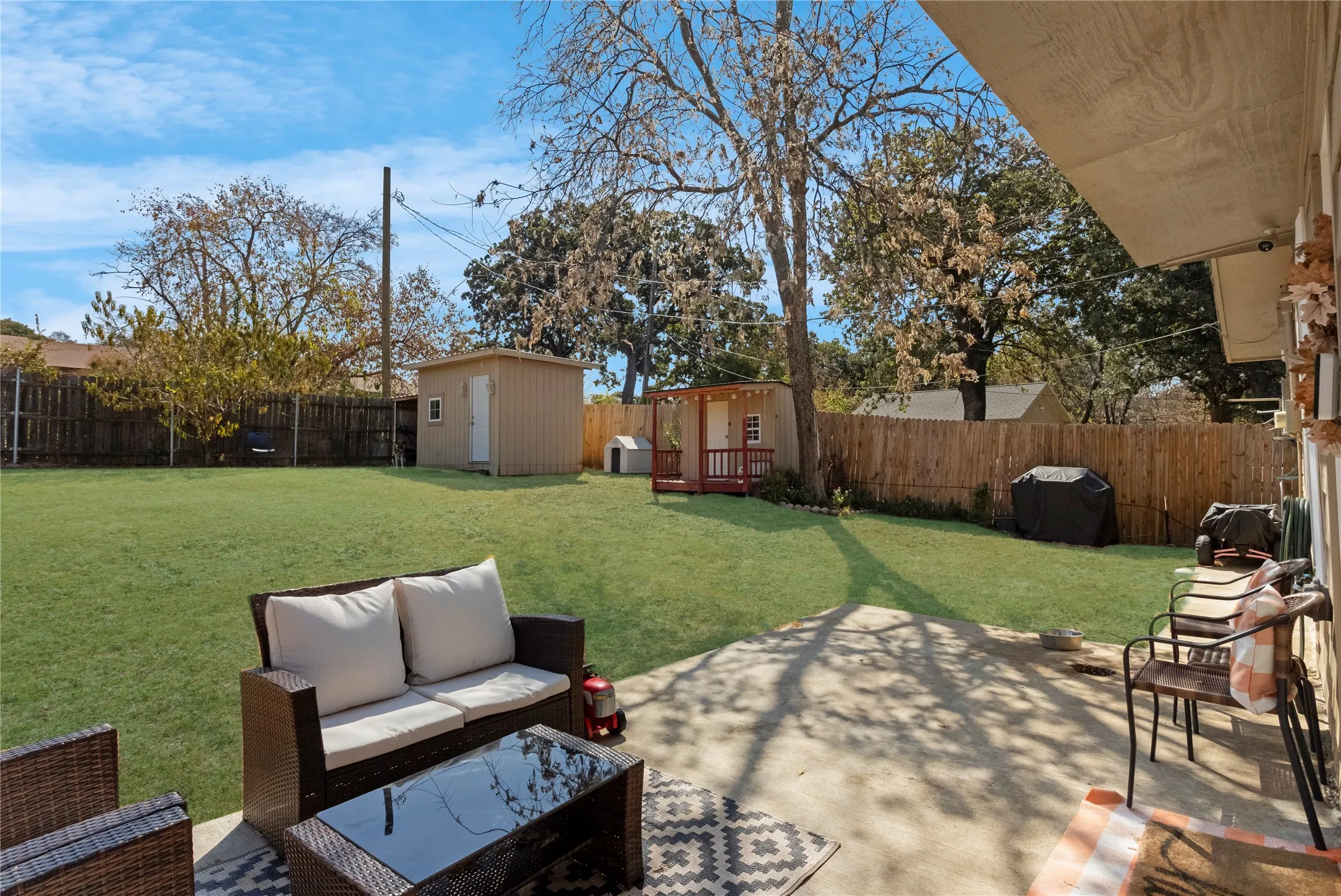 Expansive backyard featuring a concrete patio, mature trees, wood privacy fencing, and two detached storage sheds