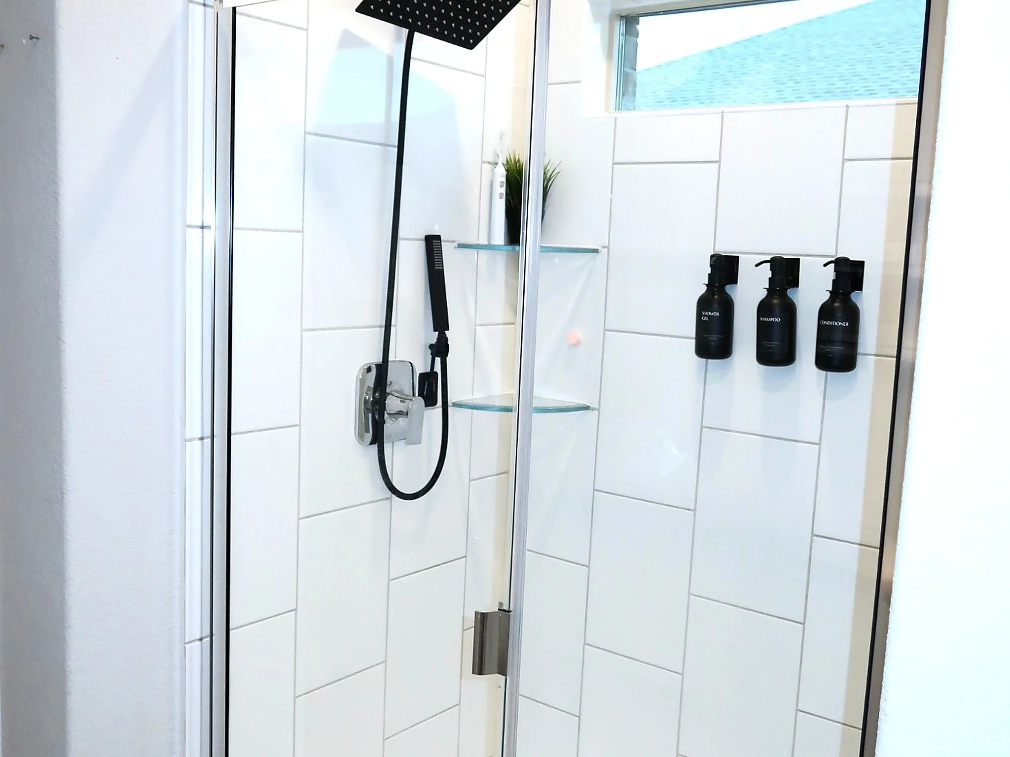 Shower enclosure with clear glass door, white rectangular tile surround, matte black rainfall shower head, and built-in glass shelving