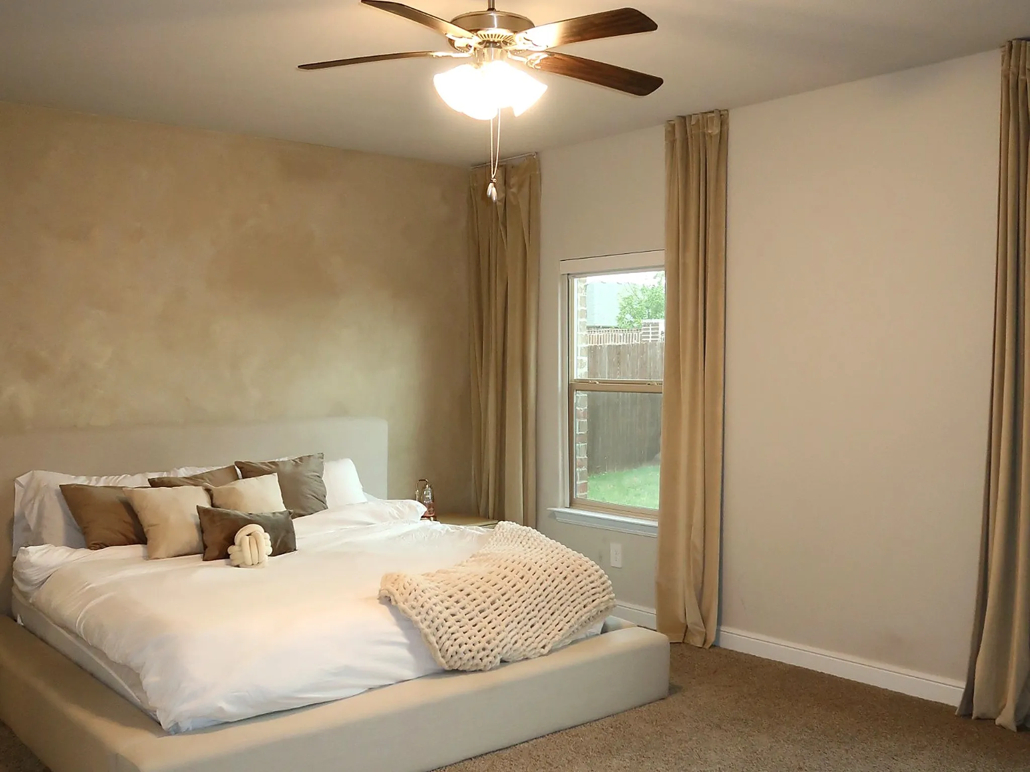 Textured accent wall, ceiling fan with integrated lighting, window with light-filtering drapery, carpeted flooring, and neutral wall finishes
