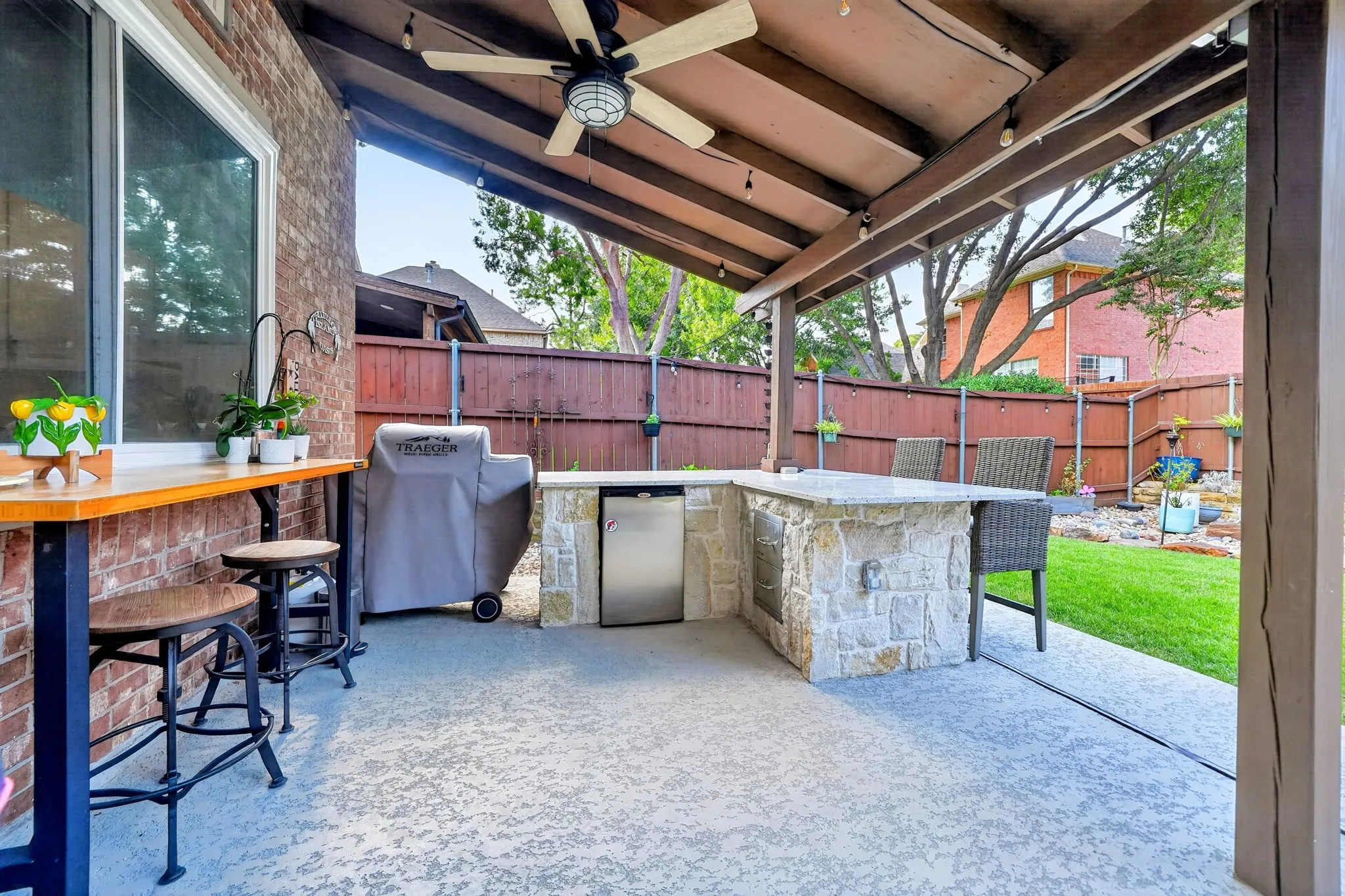 Covered patio featuring a ceiling fan, stone-clad built-in outdoor kitchen with an integrated refrigerator, concrete flooring, and a privacy fence