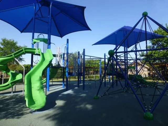 Community playground featuring blue shade structures, green slides, a climbing net, and rubberized surfacing