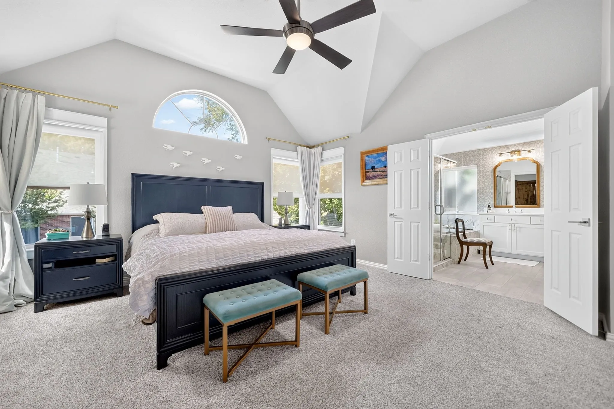 Vaulted ceiling with ceiling fan, arched transom window, dual windows with roller shades, and double doors opening to an adjoining bathroom featuring a glass-enclosed shower and dual vanity