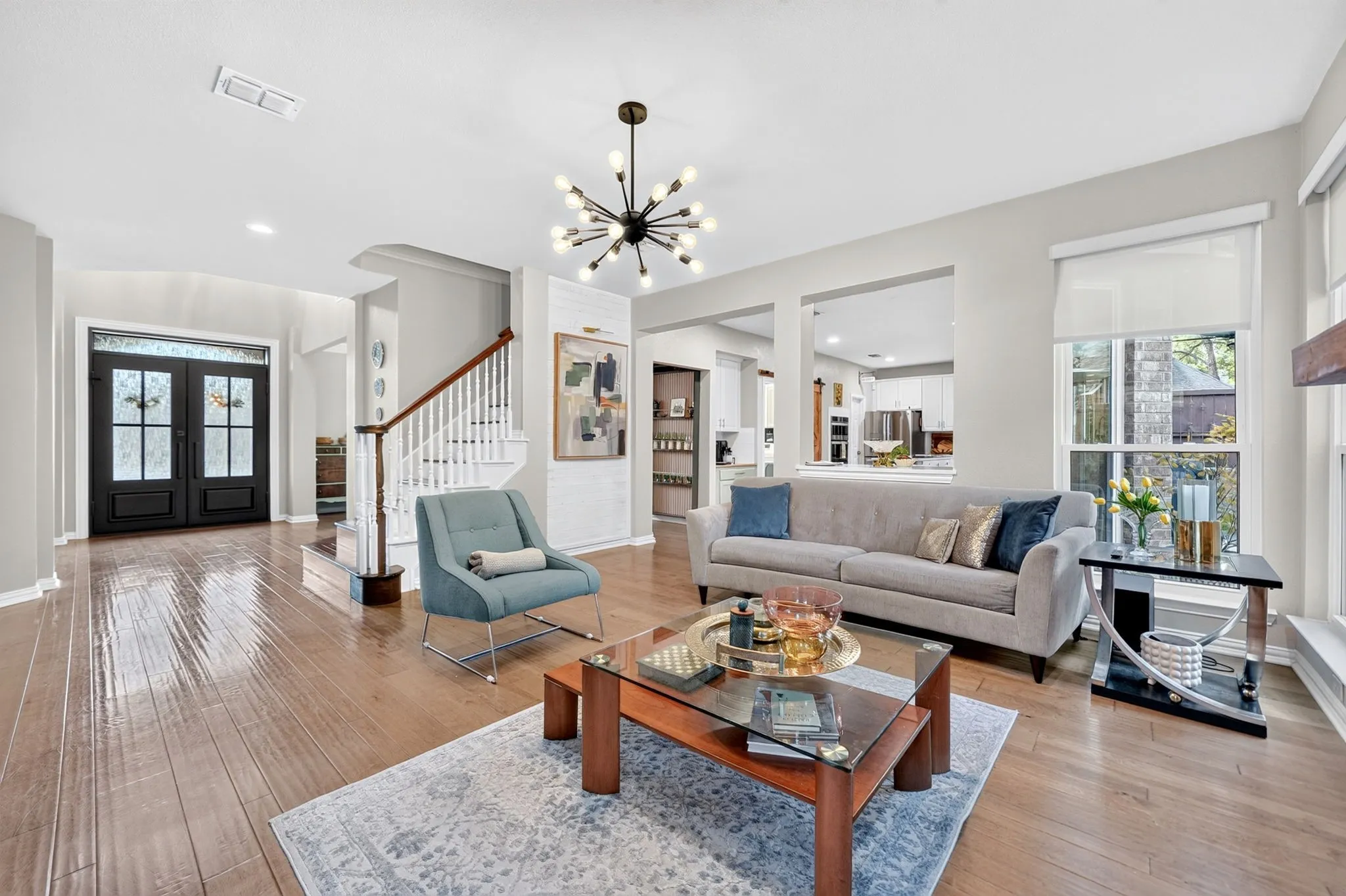 Wood-finish flooring throughout, double entry doors with decorative glass, white spindle staircase with wood handrail, and a modern sputnik chandelier