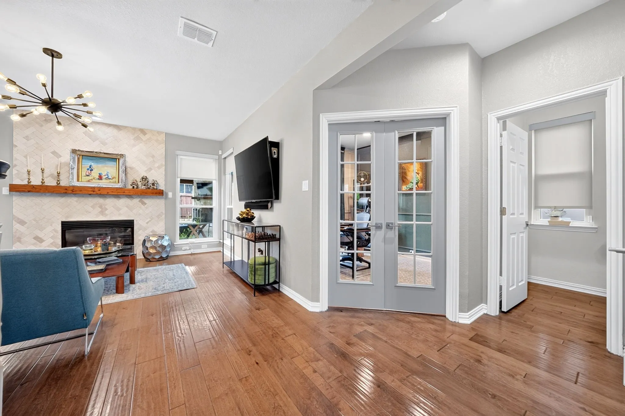 Hardwood flooring, fireplace with a herringbone tile surround, vaulted ceiling, contemporary chandelier, and French doors with glass panels