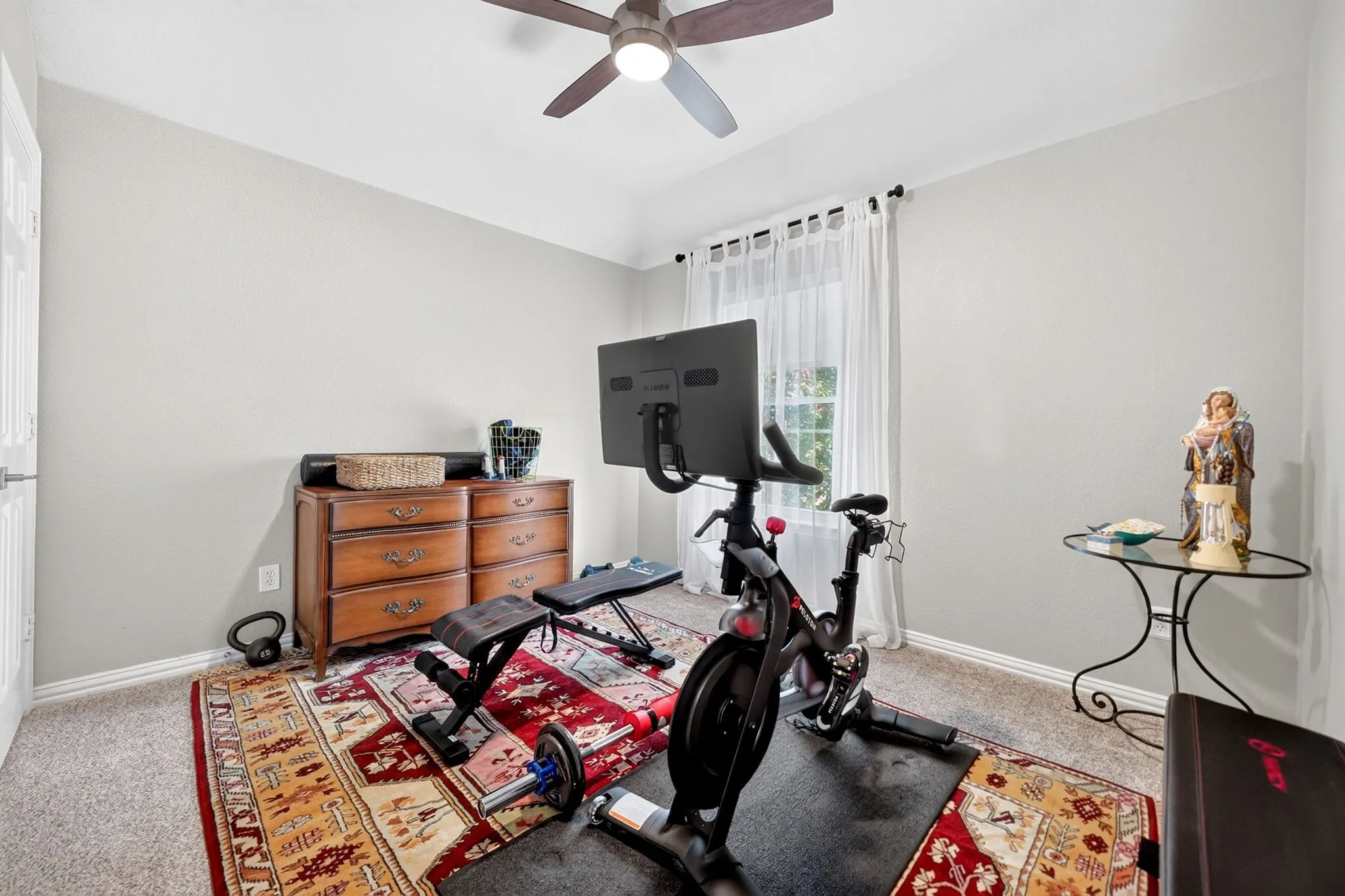 Carpeted flooring, a window with white sheers, a ceiling fan, and light-colored walls