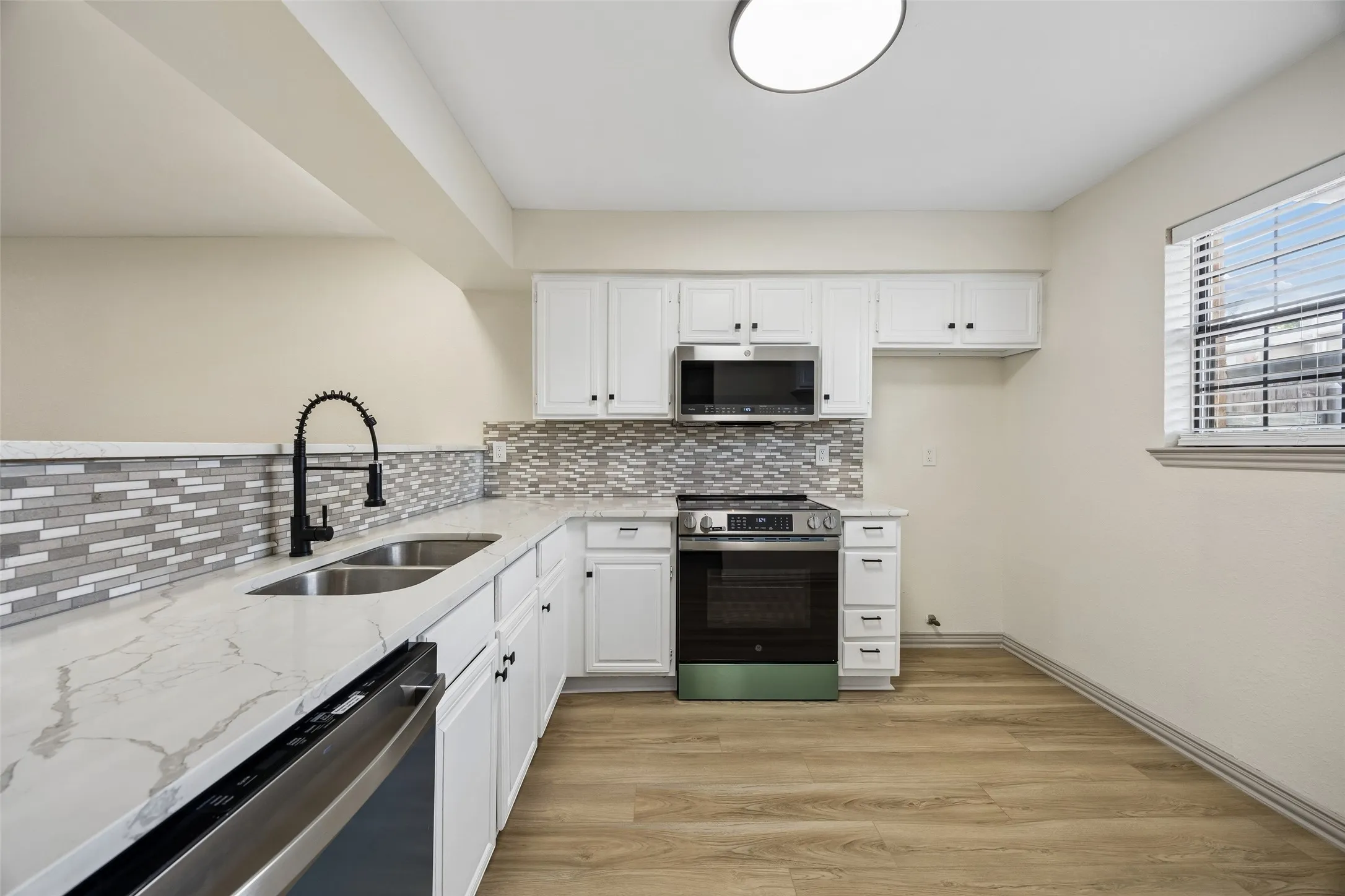 Contemporary kitchen featuring white cabinetry, a glass tile backsplash, stone-finish countertops, stainless steel appliances, and wood-finish flooring