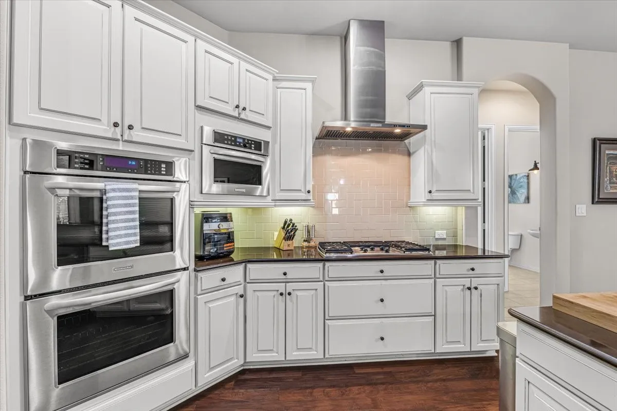 White cabinetry with dark hardware, stainless steel double ovens, a gas cooktop, and a stainless steel range hood