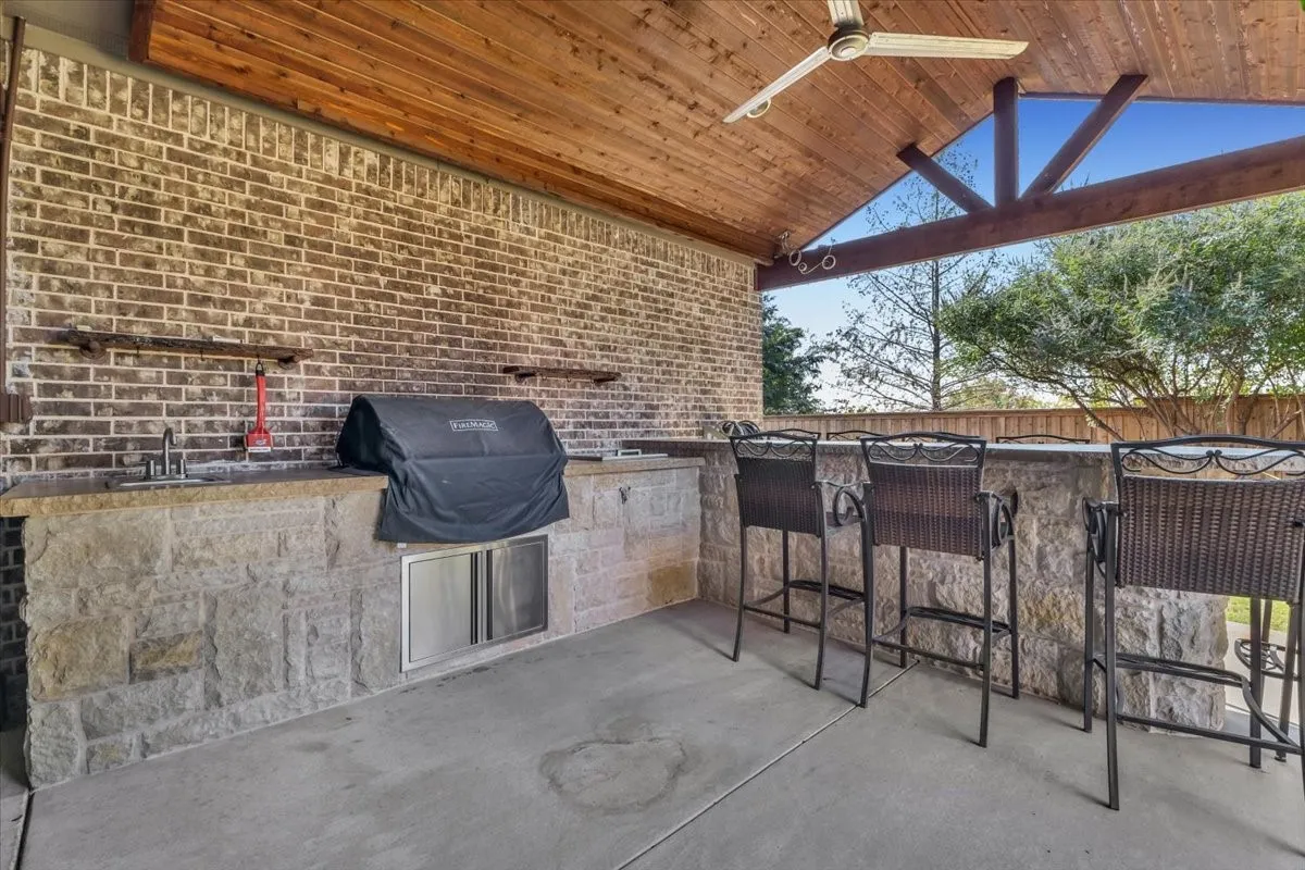 Covered outdoor kitchen featuring a wood-panel ceiling, brick wall, stone-front counter with integrated sink, and built-in grill
