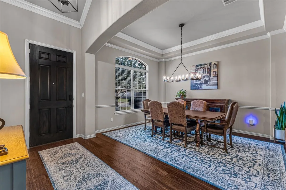Dark wood-finish flooring, arched entryway, tray ceiling with crown molding, chandelier lighting, and a large arched window with blinds
