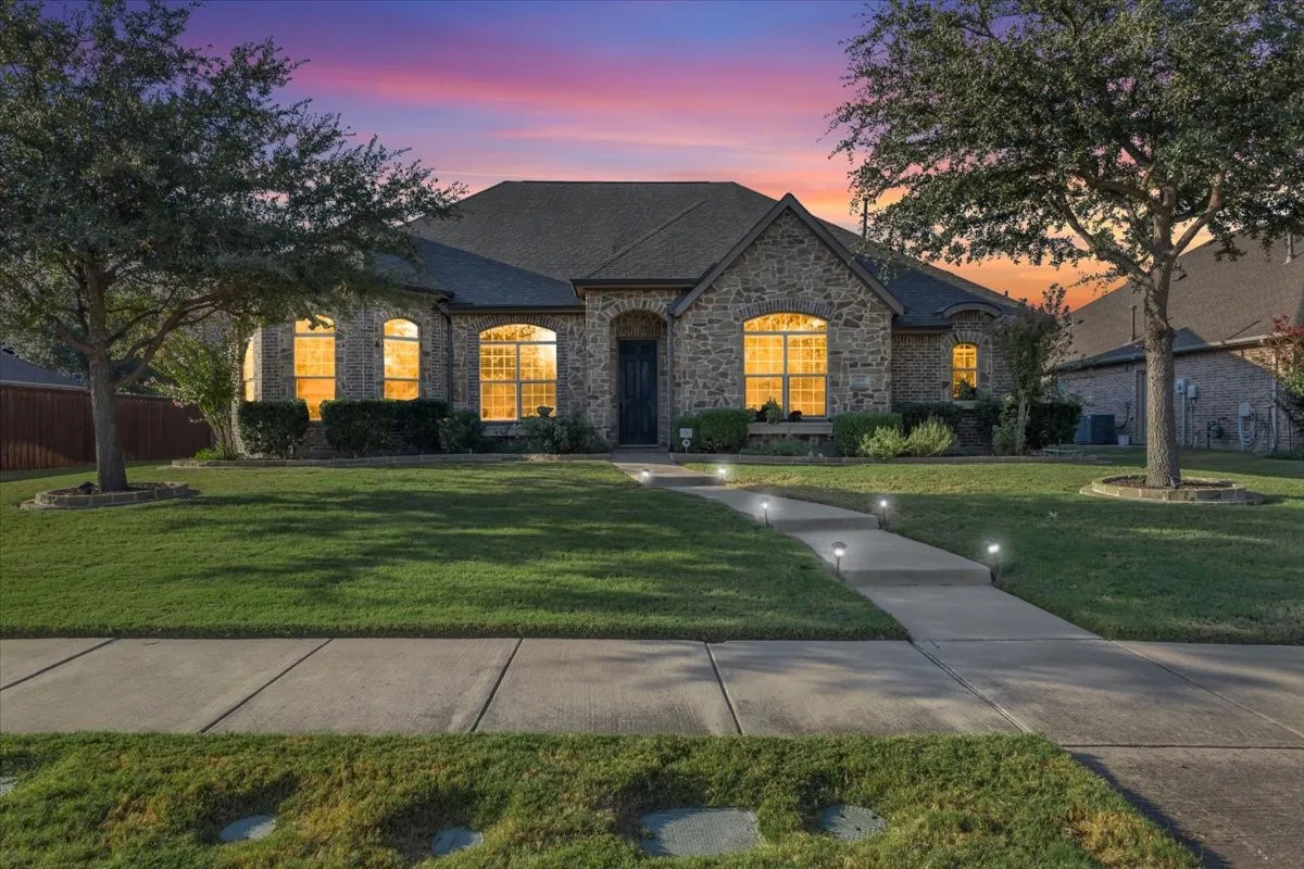 Stone facade residence with arched windows, a dark shingle roof, a concrete walkway with integrated path lighting, and a manicured lawn