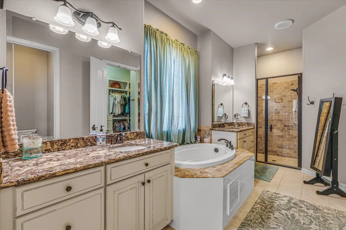 Dual vanities with stone countertops, an integrated soaking tub, a walk-in shower with glass enclosure, and a dedicated storage closet