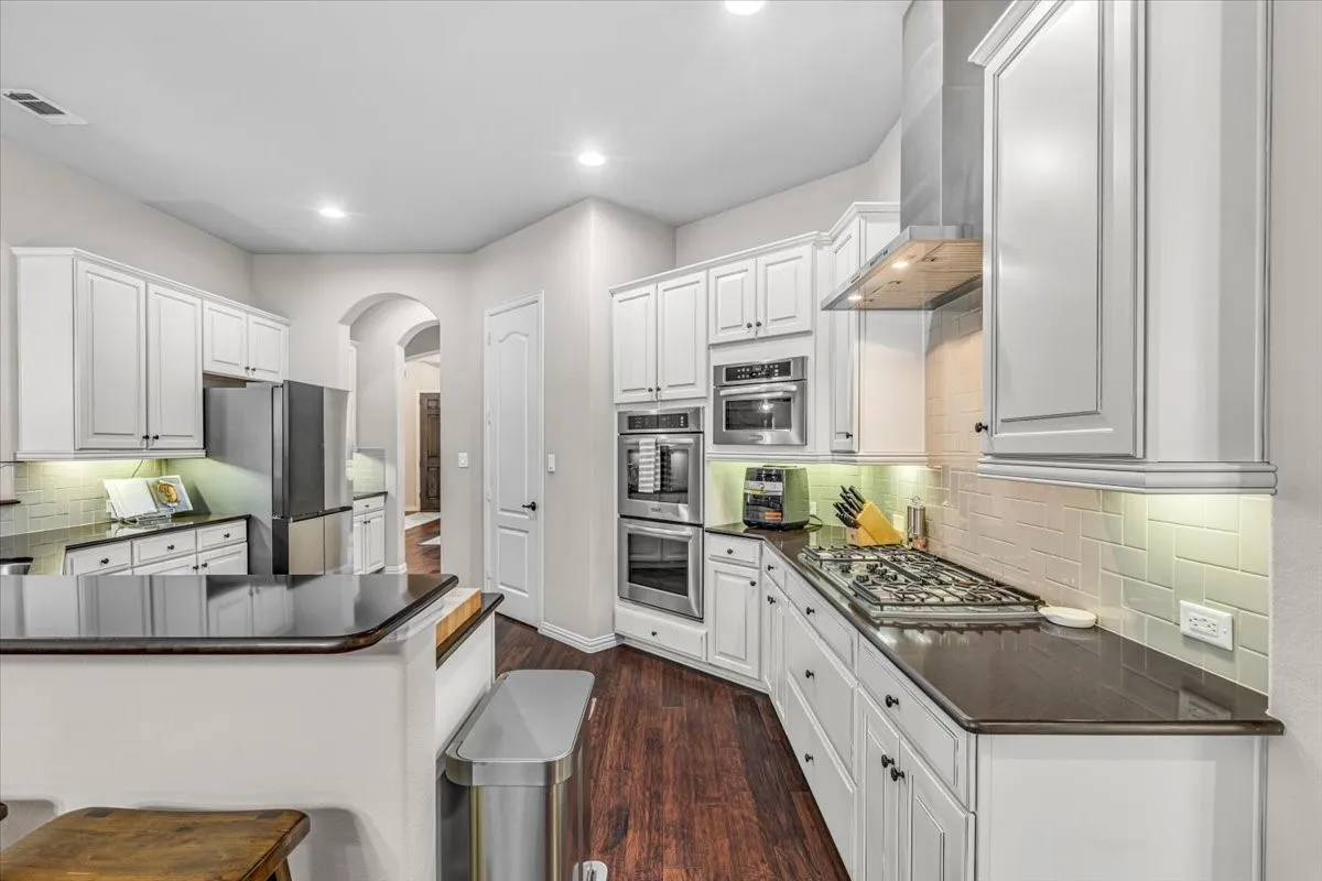 White cabinetry, dark countertops, built-in stainless steel appliances, stainless steel range hood, and wood-finish flooring
