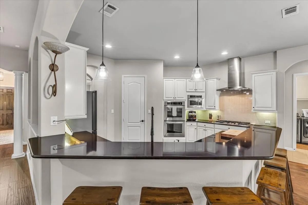Kitchen featuring white cabinetry, stainless steel appliances, a range hood, pendant lighting, and a dark countertop