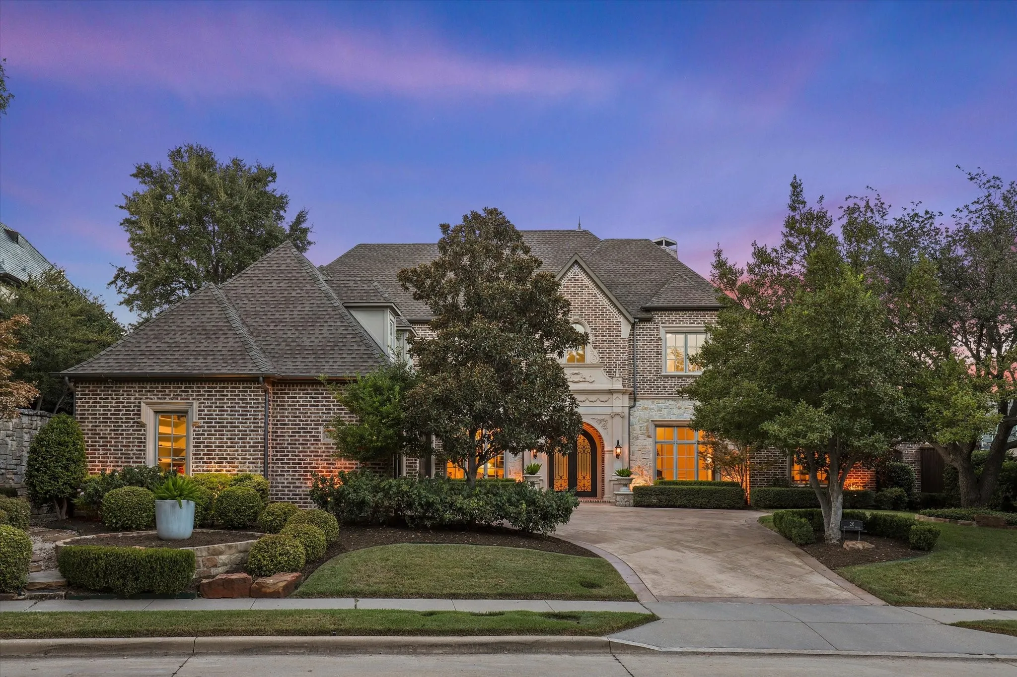 Twilight view of the timeless brick-and-stone exterior, mature trees and welcoming arched entry with custom double iron door surrounded by detailed cast stone.