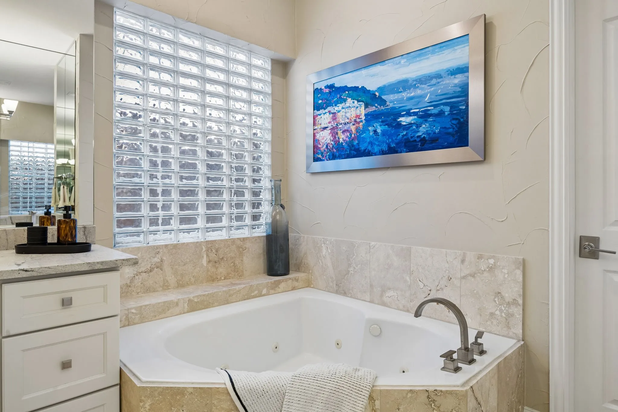 The spacious primary bathroom is light and bright, featuring travertine, separate vanities with ceiling-height mirrors and a striking glass block window that creates a space full of natural light.