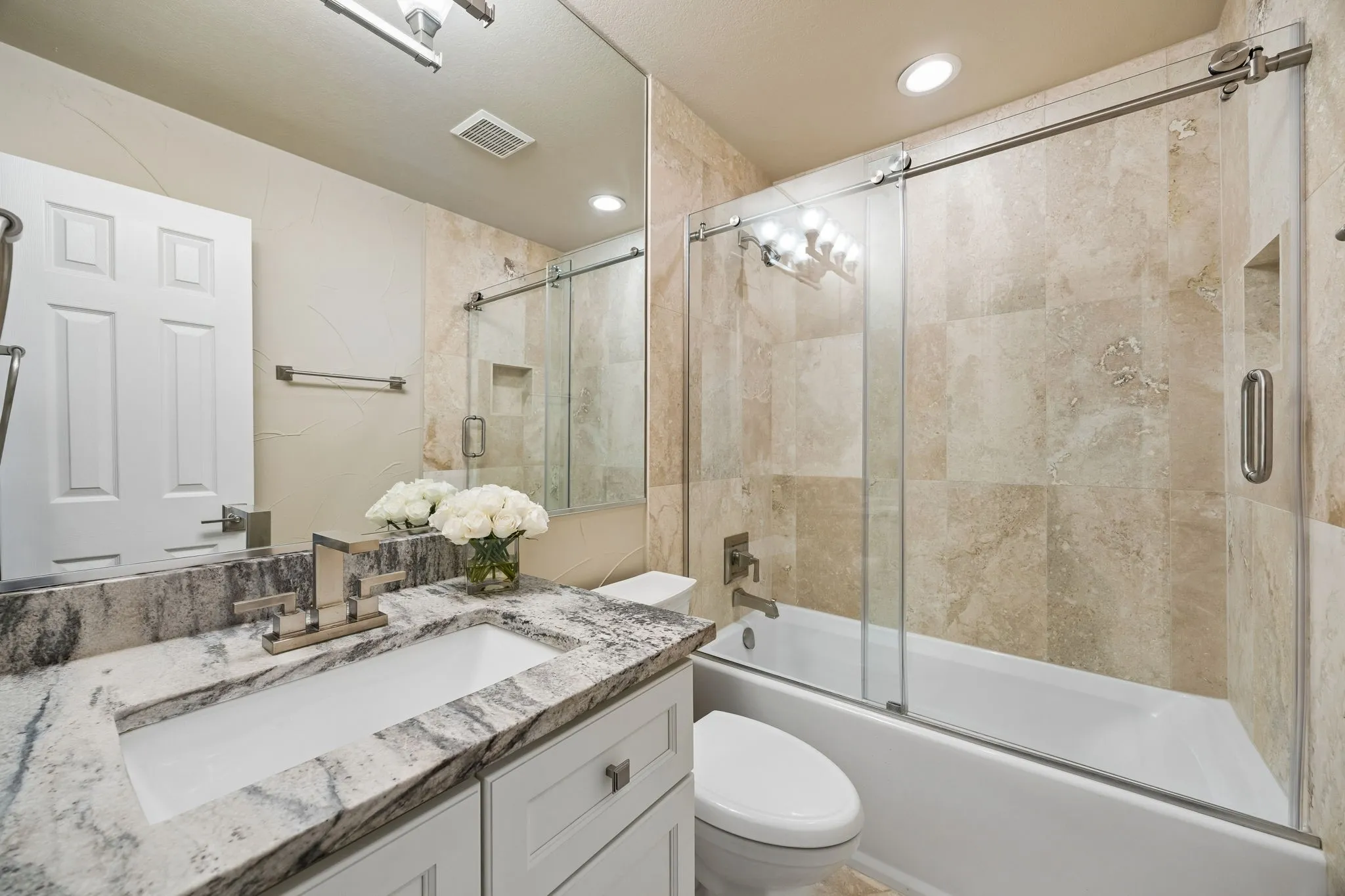 The upstairs hall bathroom features a natural stone countertop, travertine tile, and a tub/shower combination with sliding glass enclosure.  Modern fixtures, recessed lighting and ample vanity storage create a polished and comfortable space for family and guests.
