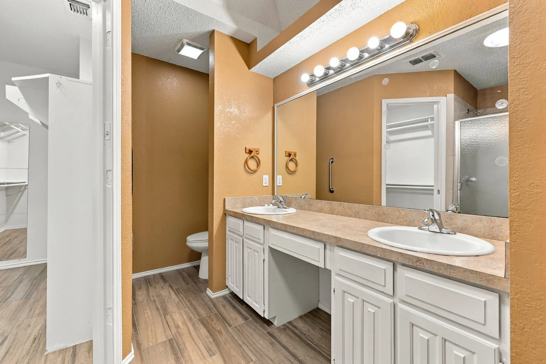 Dual sinks set in a neutral countertop with white cabinetry, wood-finish flooring, a wall-mounted vanity light fixture, and a glass-enclosed shower