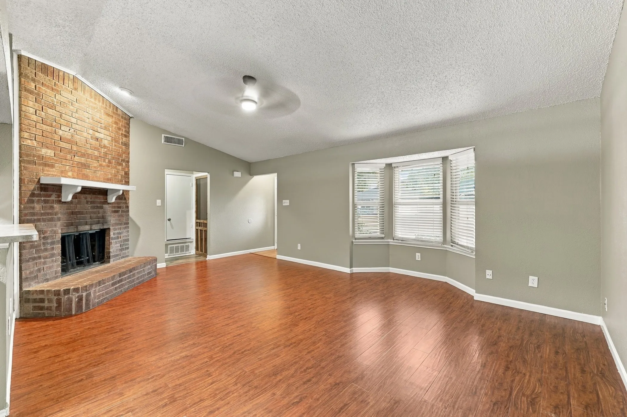 Brick fireplace with a white mantel, wood-finish flooring, bay window with blinds, vaulted ceiling, and ceiling fan