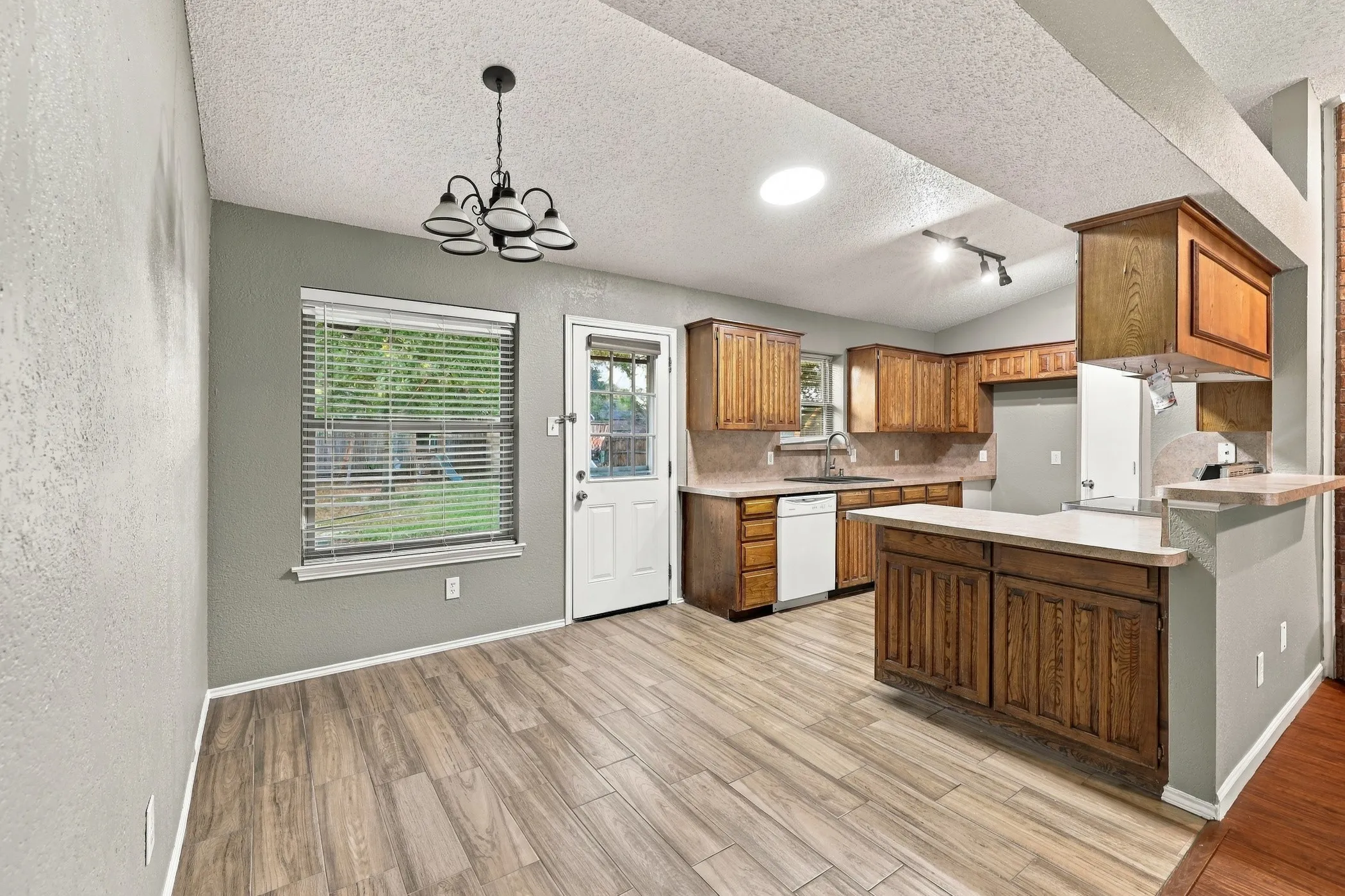 Wood-finish flooring, overhead chandelier, kitchen with wood cabinetry, light-colored countertops, and a breakfast bar