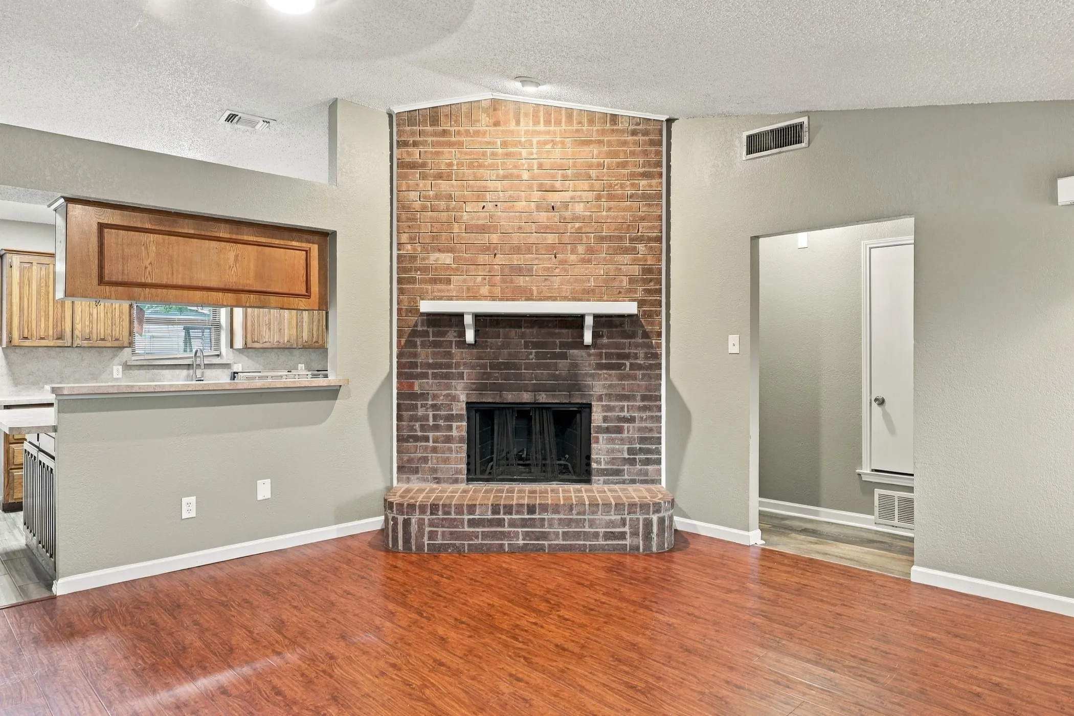 Wood-finish flooring, brick fireplace with a white mantel, open pass-through to kitchen, neutral wall tones, and recessed lighting