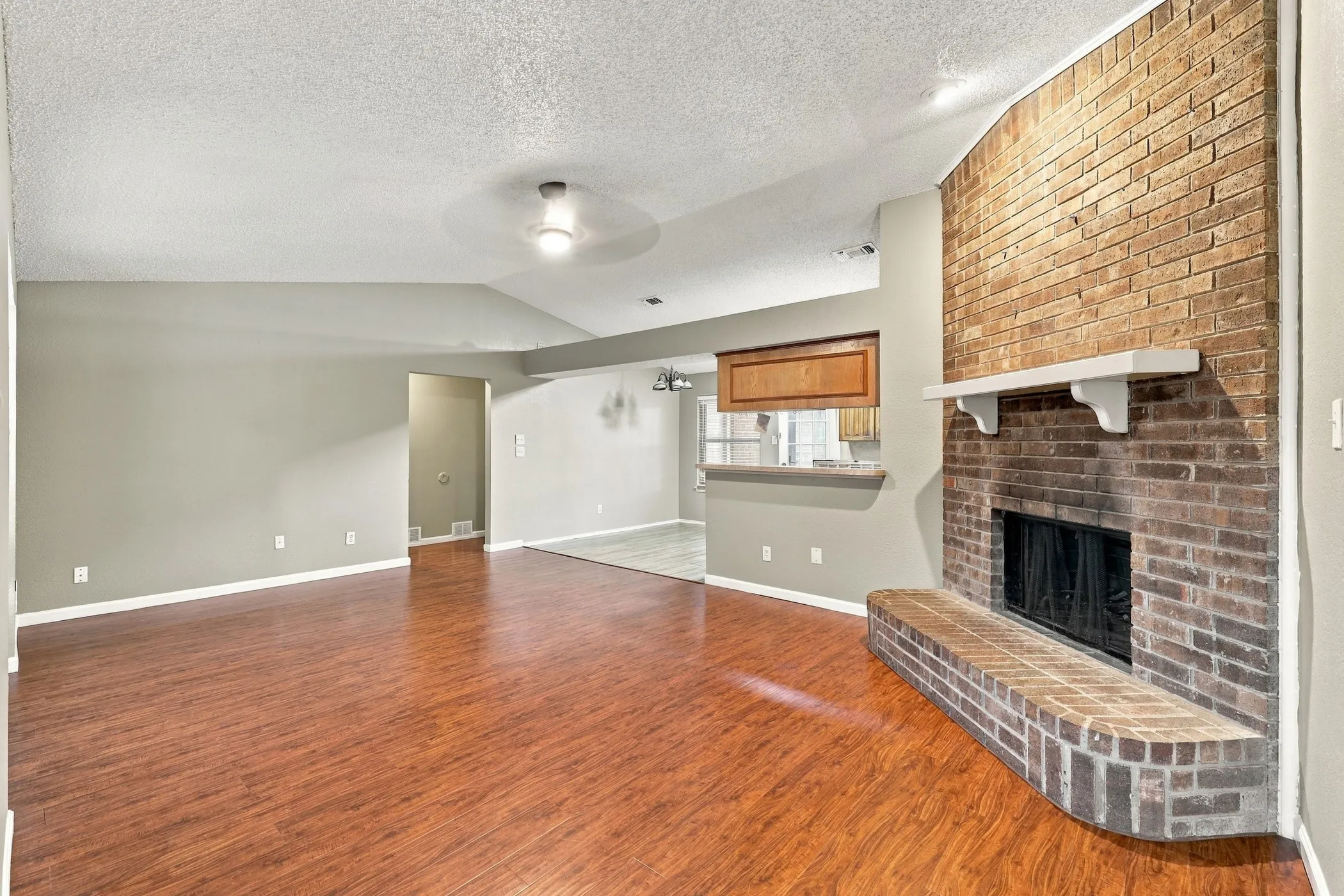 Spacious living area featuring wood-finish flooring, a vaulted ceiling, and a brick fireplace with a hearth and mantel