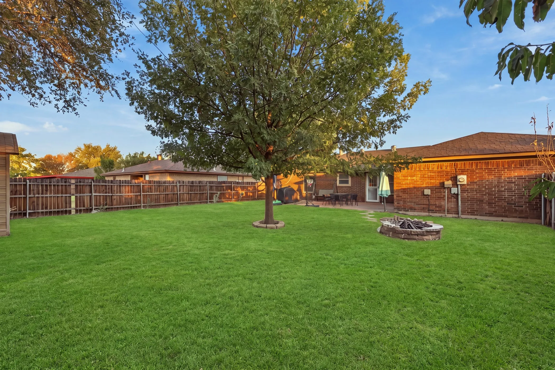 Expansive backyard featuring a manicured lawn, a mature tree, a built-in stone fire pit, a wood privacy fence, and a brick exterior