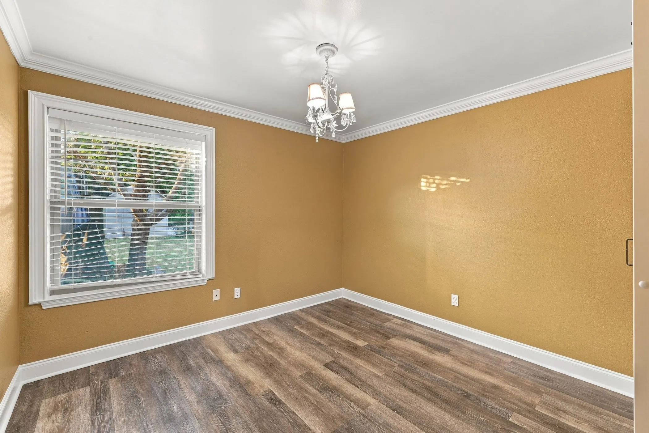 Wood-finish flooring, white crown molding, a window with white blinds, and a white chandelier with sconce lighting