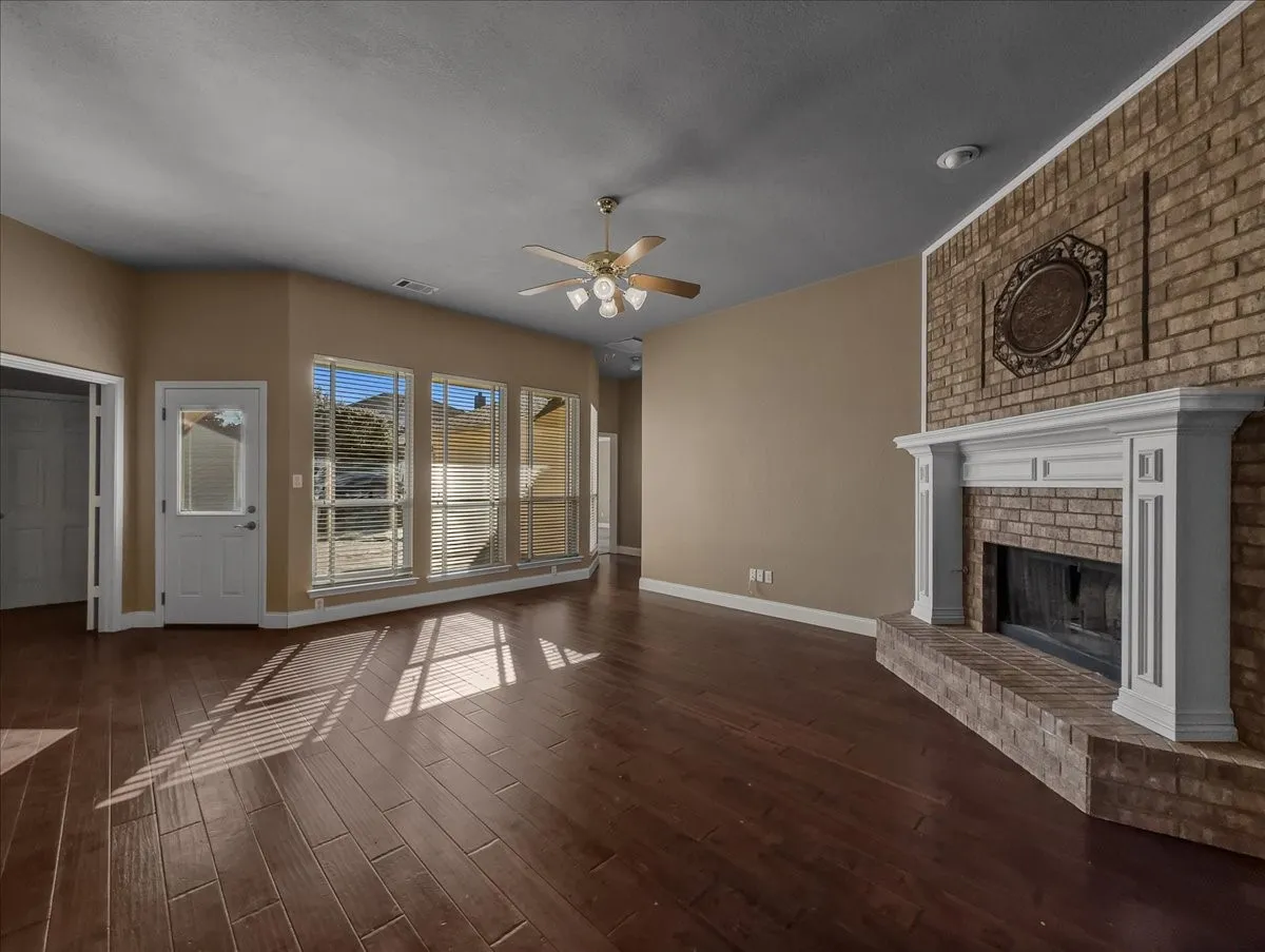 Brick fireplace with a white mantel, wood-finish flooring, multiple windows with horizontal blinds, a ceiling fan with integrated lighting, and neutral wall tones