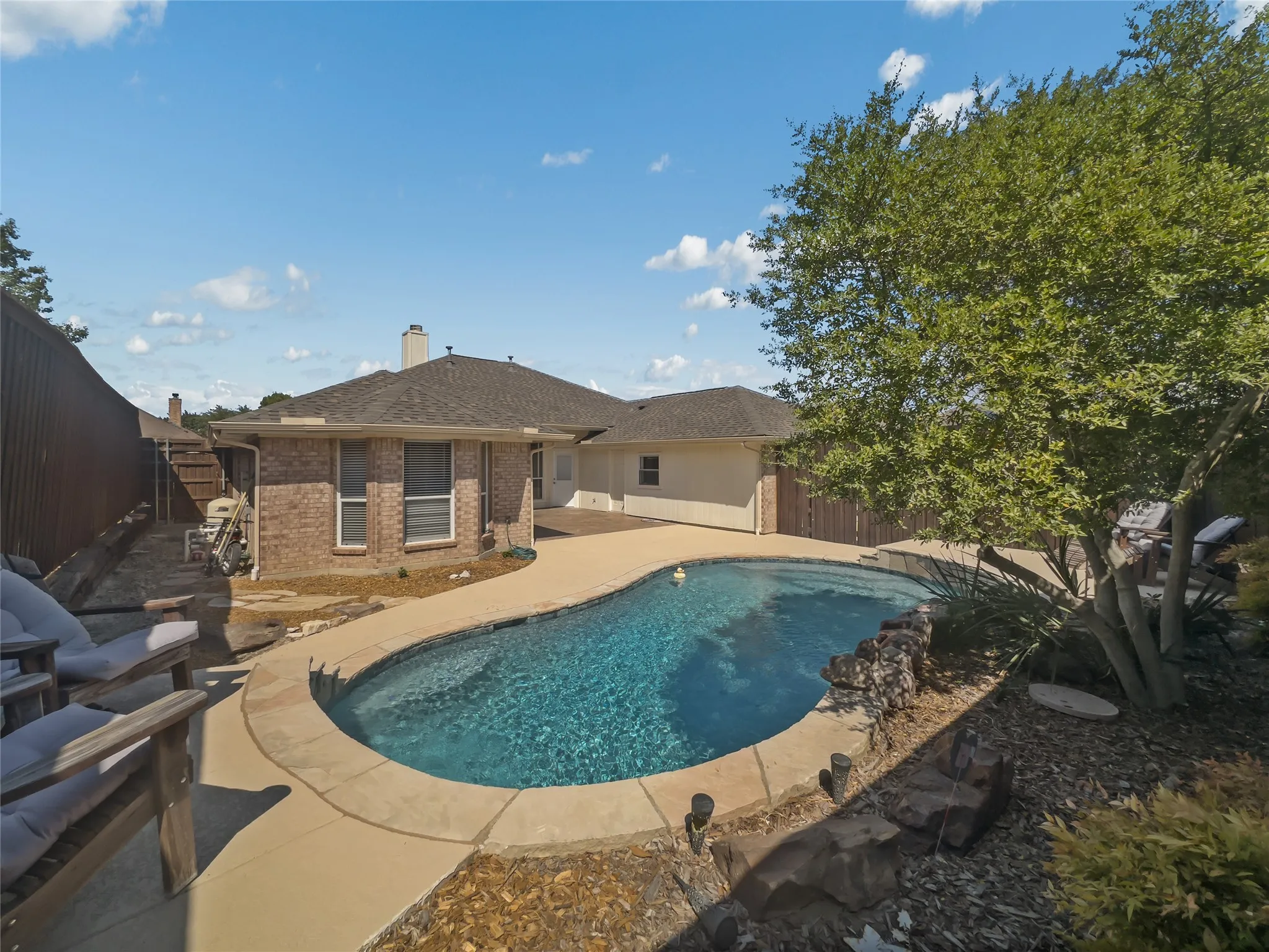 Backyard swimming pool with paved surround, brick exterior, and mature tree