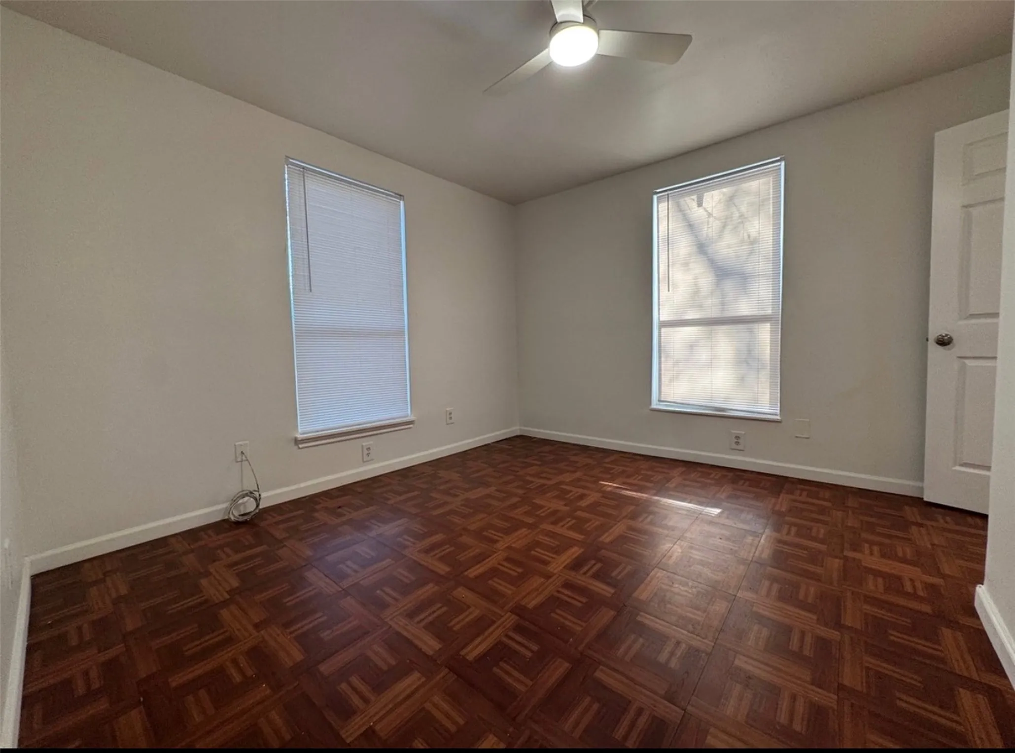 Hardwood parquet flooring with white baseboards, a ceiling fan with integrated lighting, and two windows with blinds