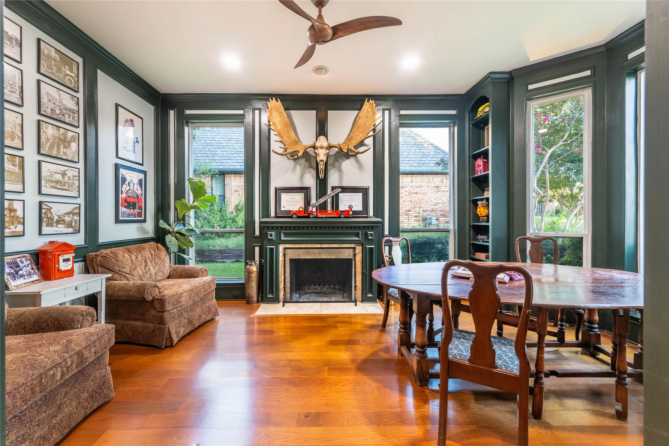 Hardwood flooring, fireplace with stone surround, built-in shelving, ceiling fan, and large windows