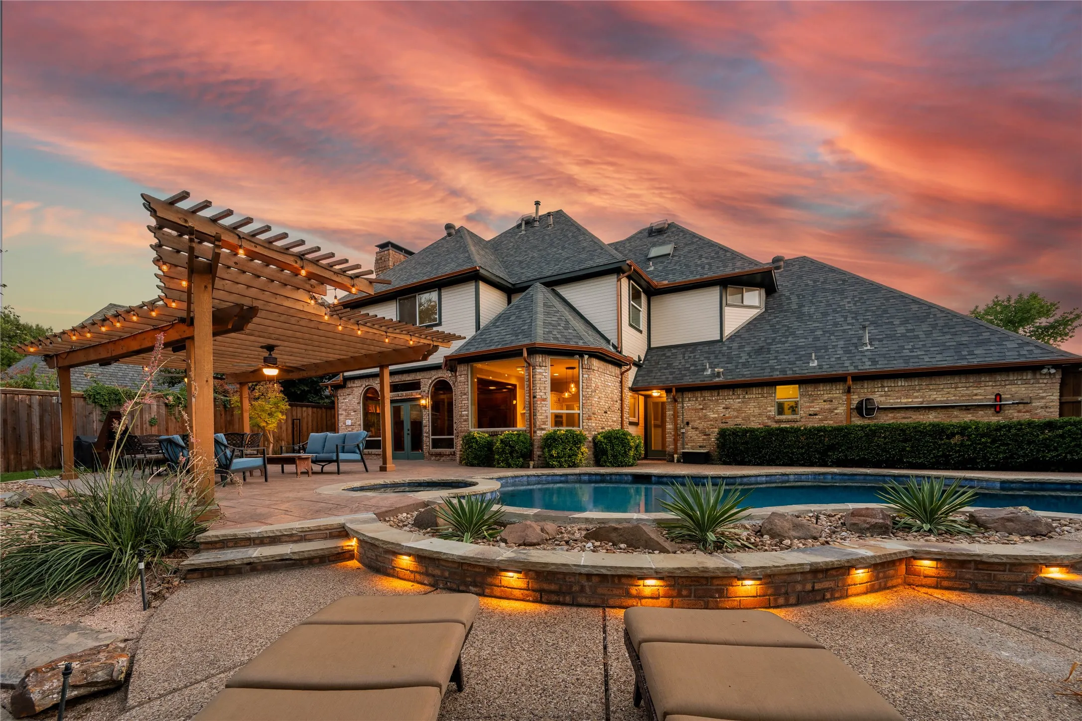 Expansive outdoor living area featuring a custom wood pergola, built-in swimming pool, integrated spa, and illuminated landscaping with stone accents