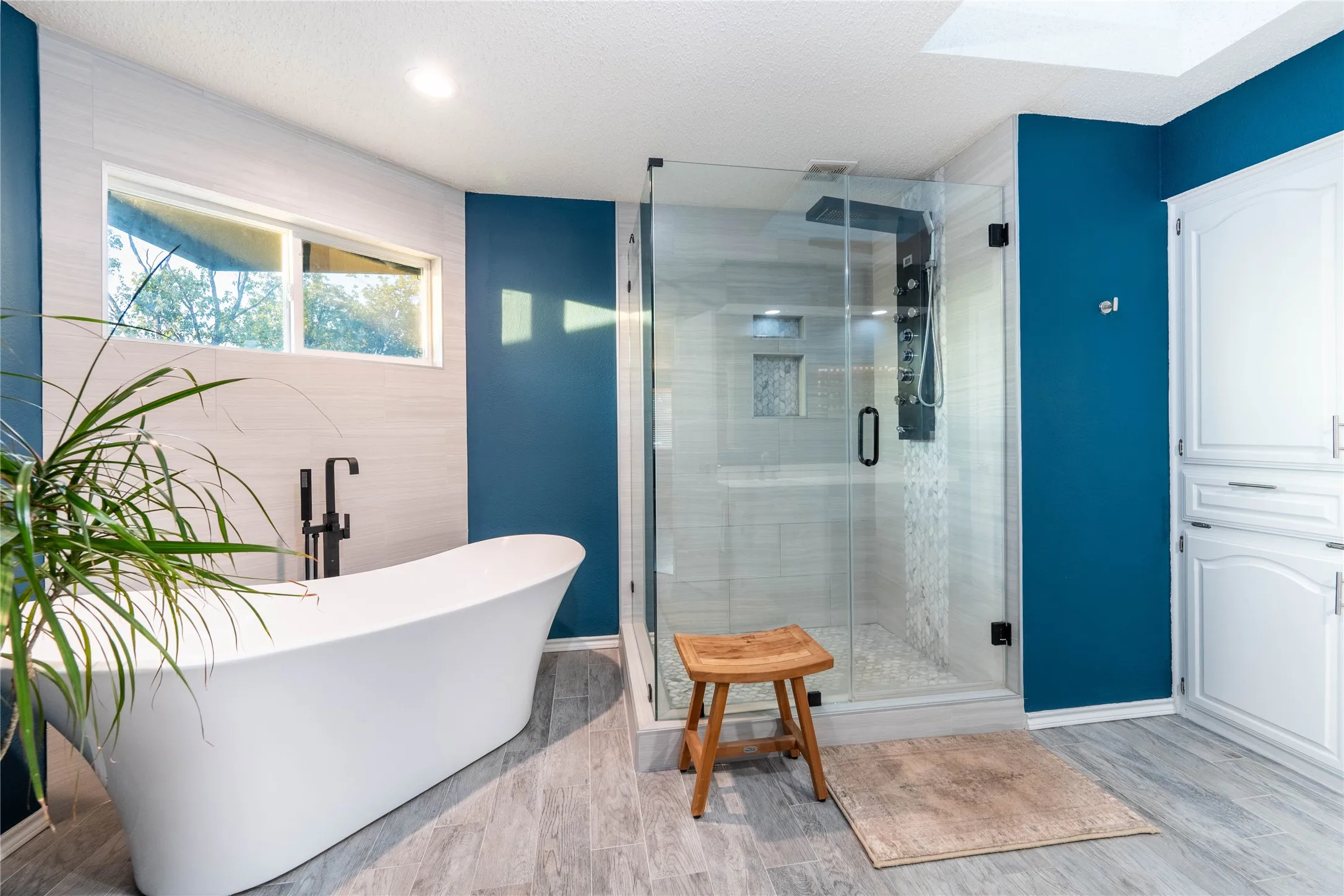 Freestanding bathtub with a floor-mounted matte black faucet, glass-enclosed shower with a multi-function panel, built-in cabinetry, wood-finish flooring, and a skylight