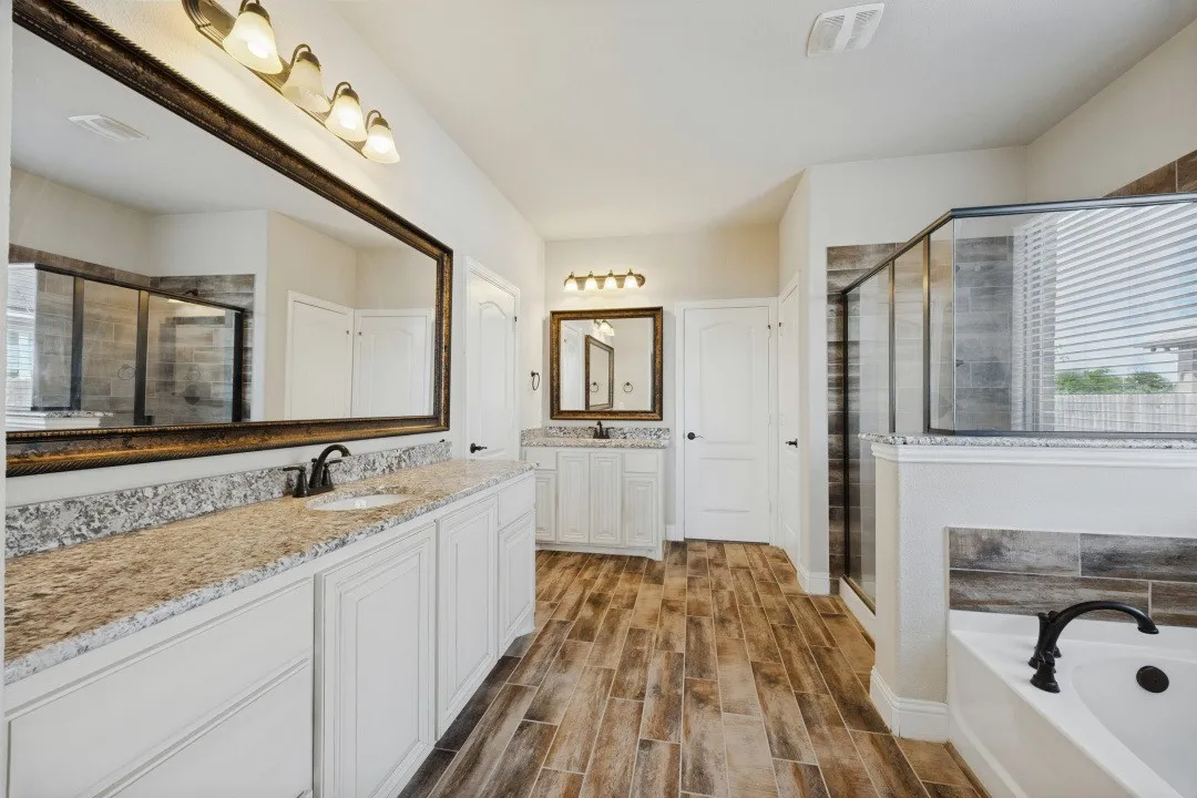 Dual vanity sinks with stone countertops, wood-finish flooring, a glass-enclosed shower, a deep soaking tub, and substantial wall mirrors