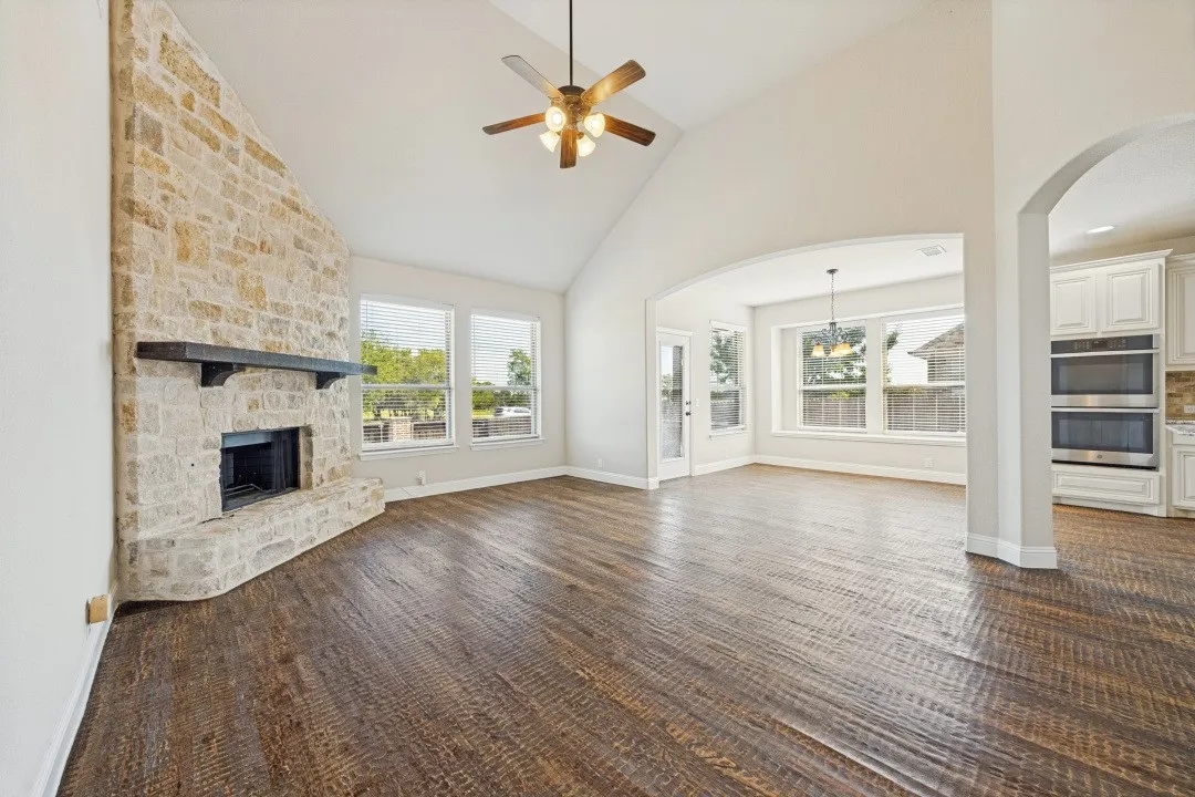 Stone-clad fireplace with mantel, vaulted ceiling with ceiling fan, wood-finish flooring, multiple windows, and arched entryway