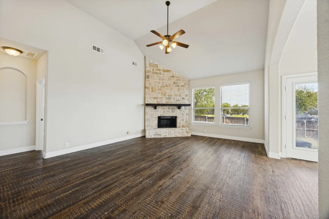 Vaulted ceiling with a ceiling fan, stone-clad fireplace, wood-finish flooring, and multiple windows