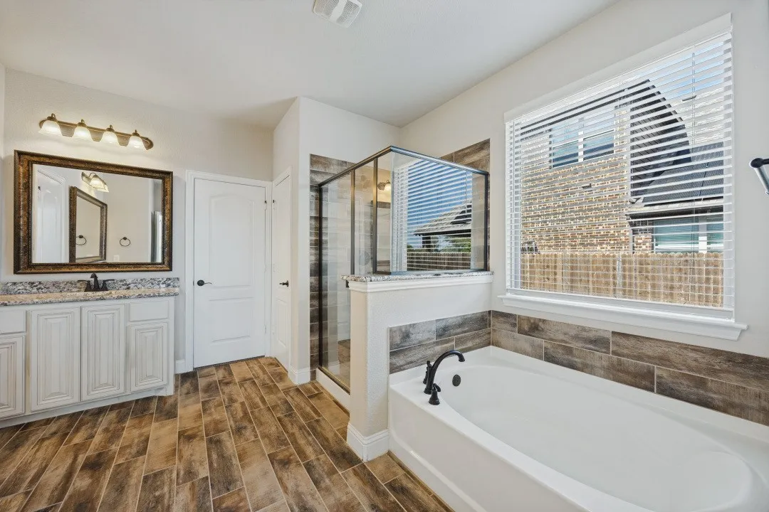 Wood-finish flooring, white vanity with granite-style countertop, large framed mirror, glass-enclosed shower, and a built-in soaking tub