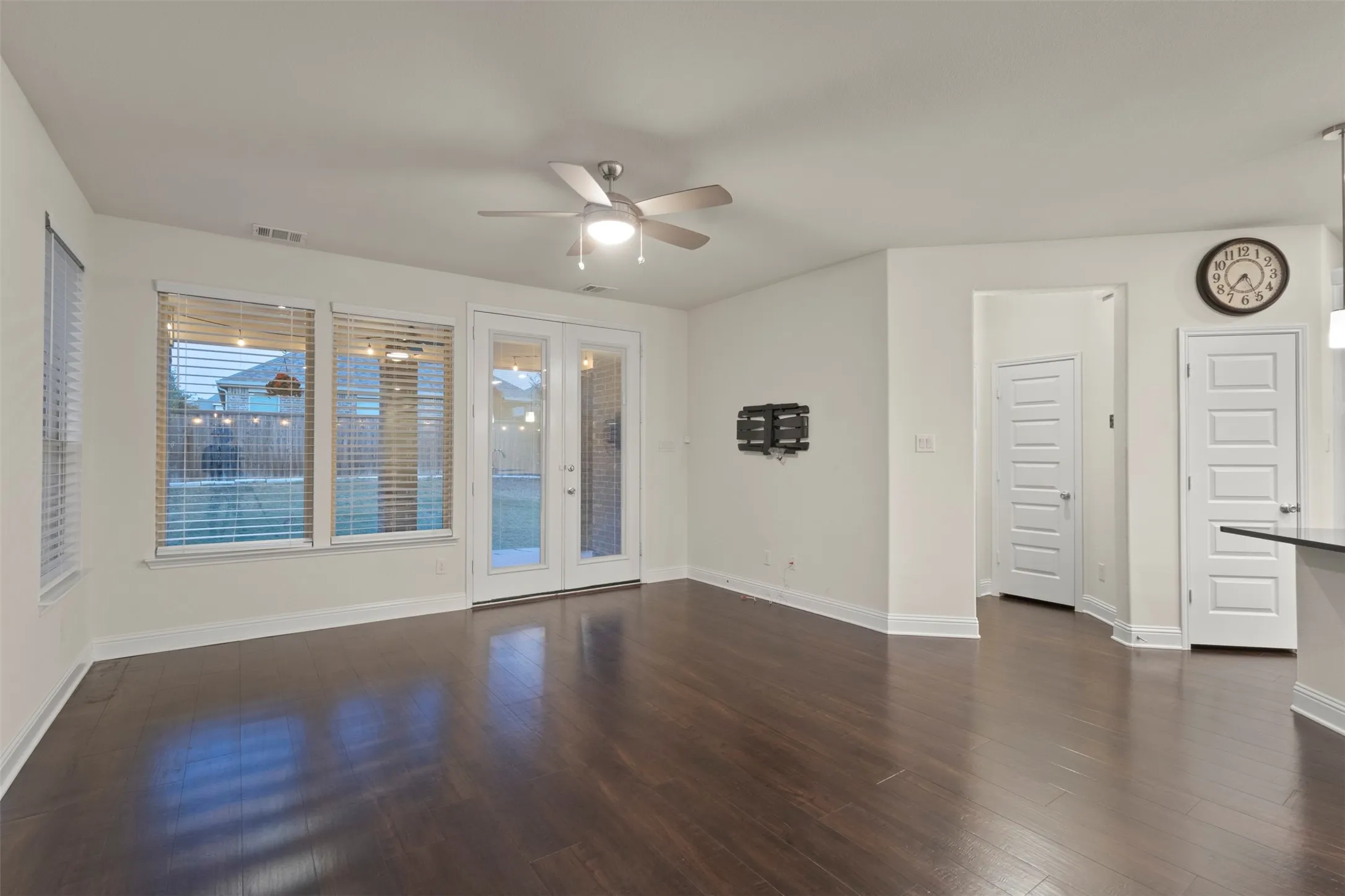 Dark wood-finish flooring, white walls, ceiling fan with integrated lighting, glass-paned double doors, and multiple windows
