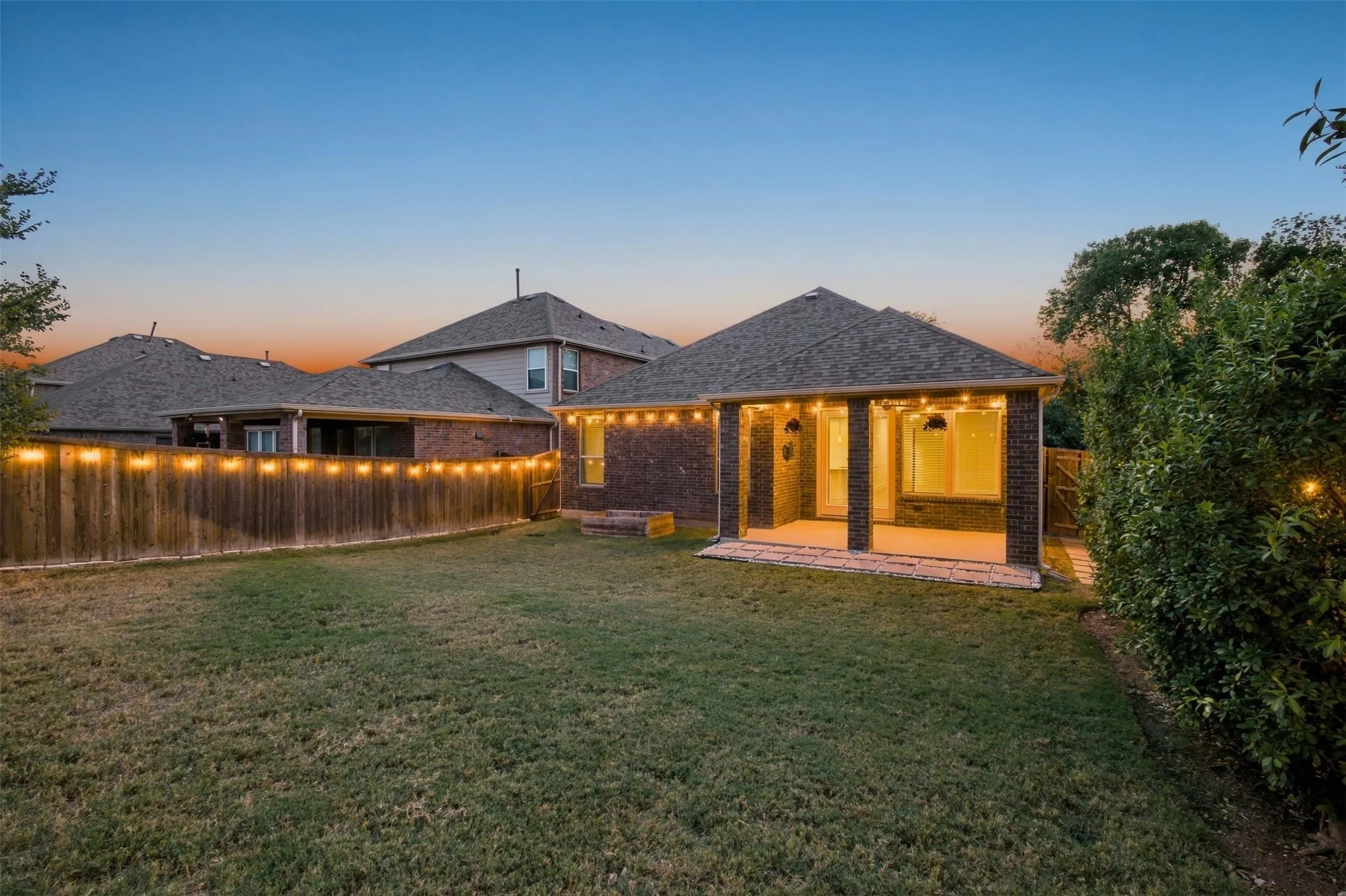 Expansive backyard featuring a covered patio with brick columns, string lighting, and a wood privacy fence