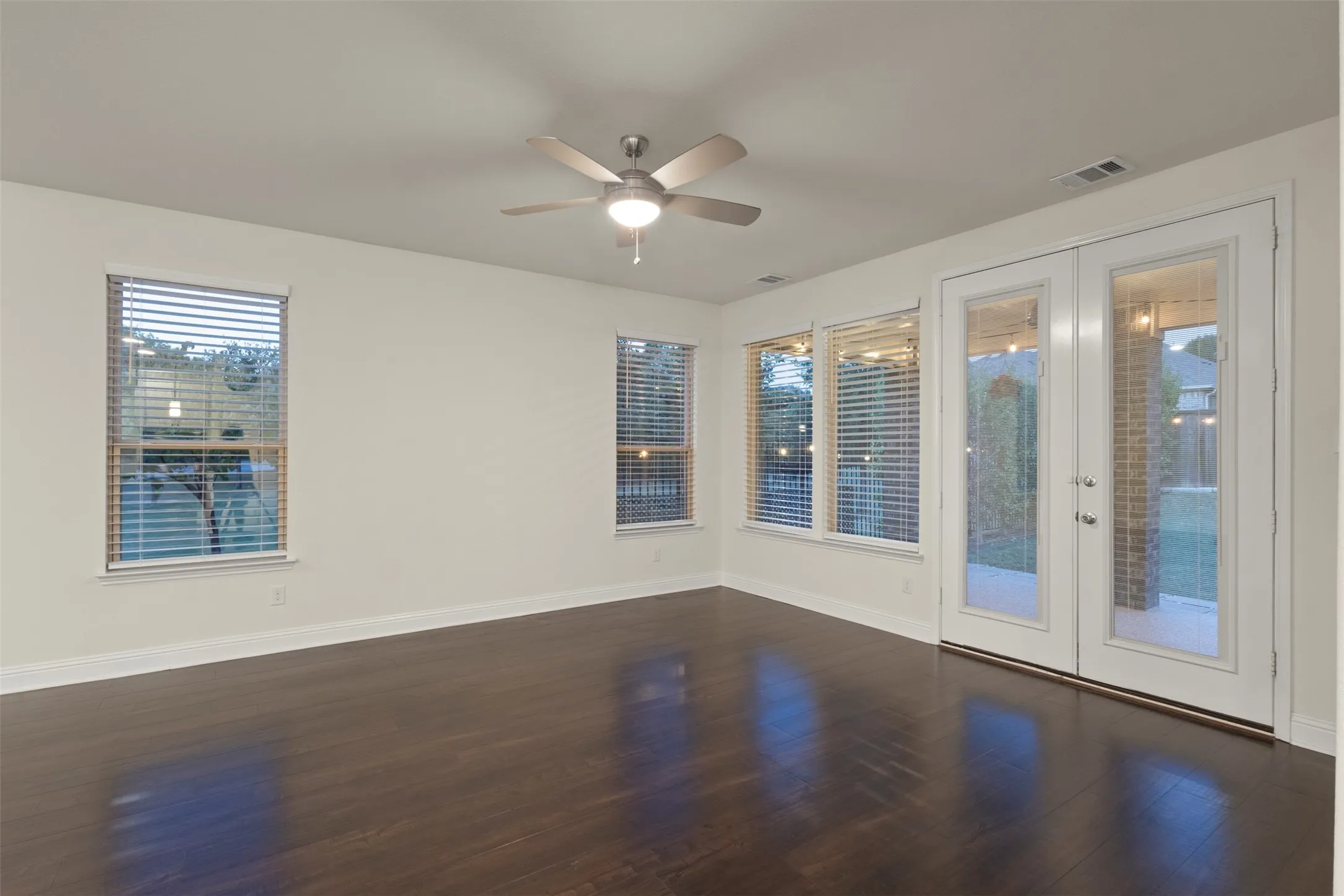 Dark wood-finish flooring, white walls, multiple windows with blinds, double glass-paneled doors, and a ceiling fan with integrated lighting