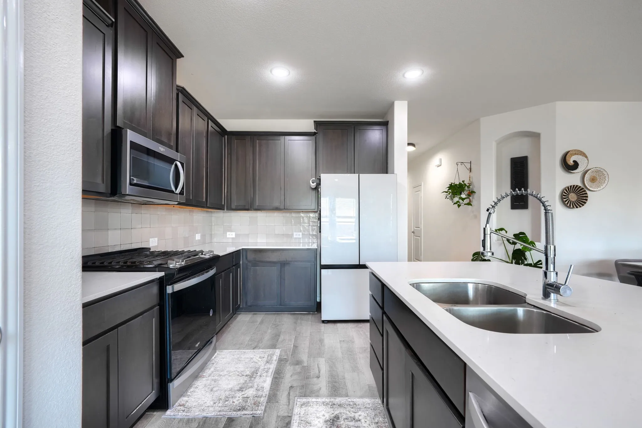 Kitchen featuring dark wood-finish cabinetry, light-toned countertops, a stainless steel double basin sink with a high-arch faucet, a built-in microwave, and wood-finish flooring