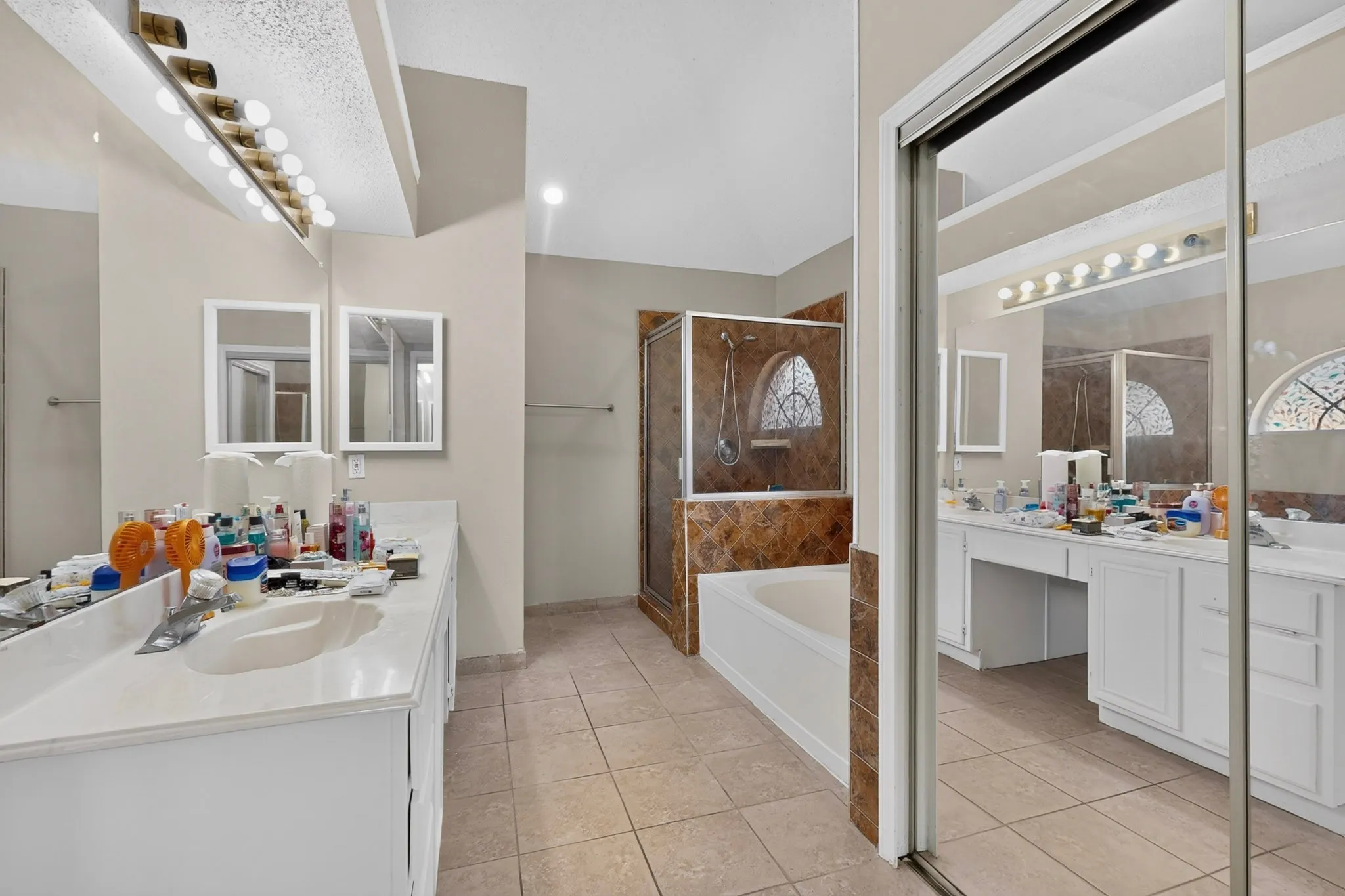 Dual vanity with integrated sinks, full-width mirror, tile flooring, built-in bathtub, and a separate shower with glass enclosure