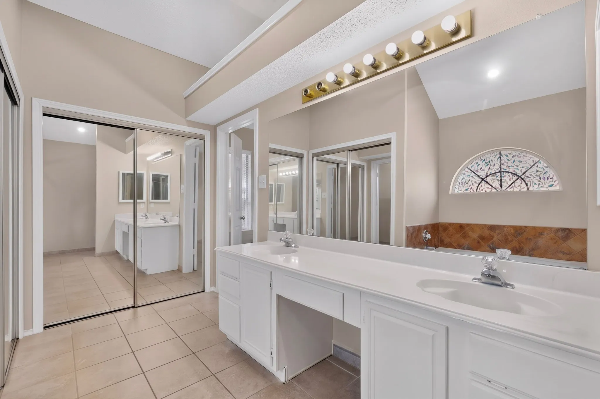 Dual vanities with white cabinetry, tile flooring, mirrored sliding closet doors, and vaulted ceiling