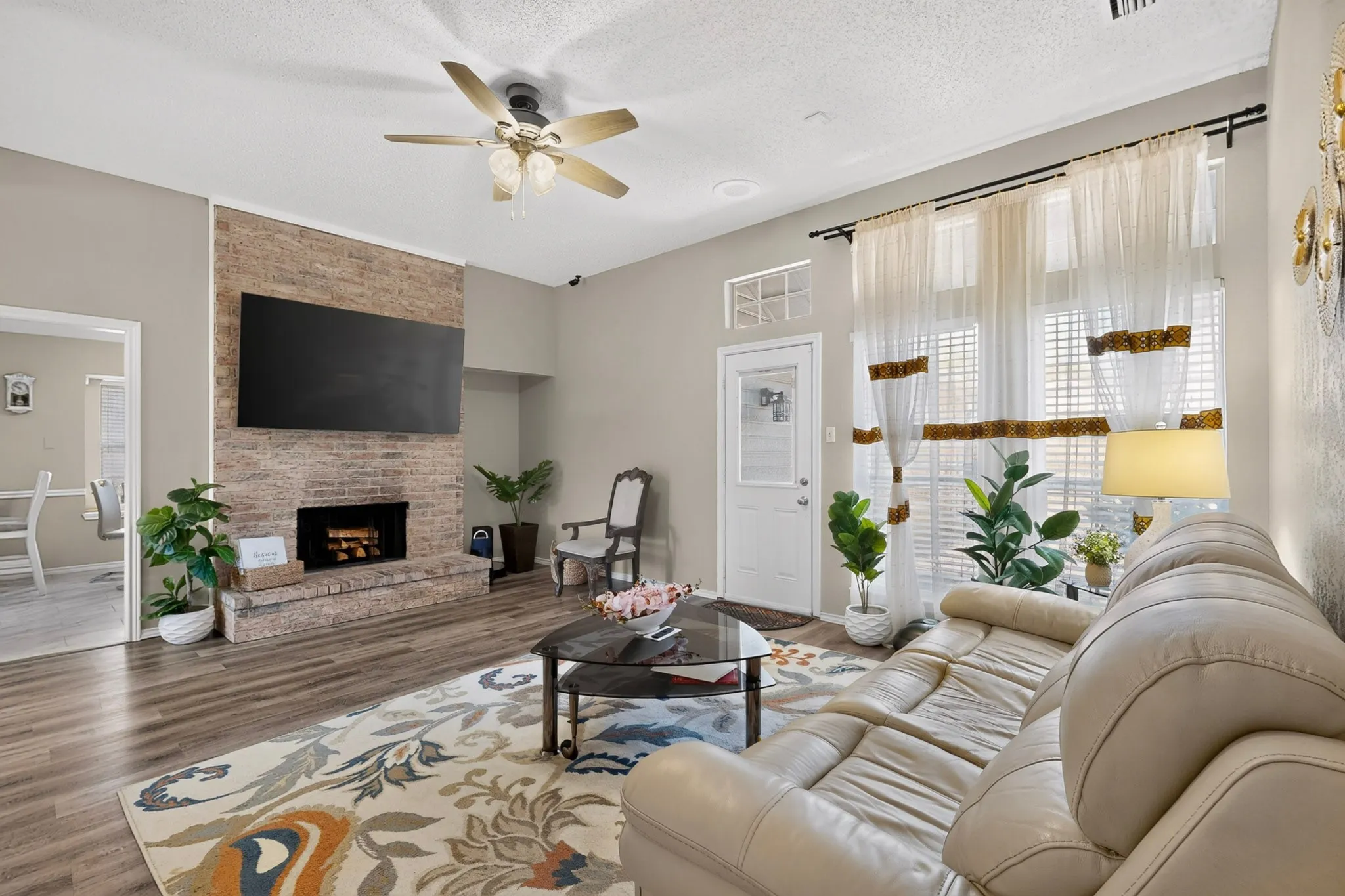 Spacious living area featuring a brick fireplace, wood-finish flooring, and ample natural light from large windows