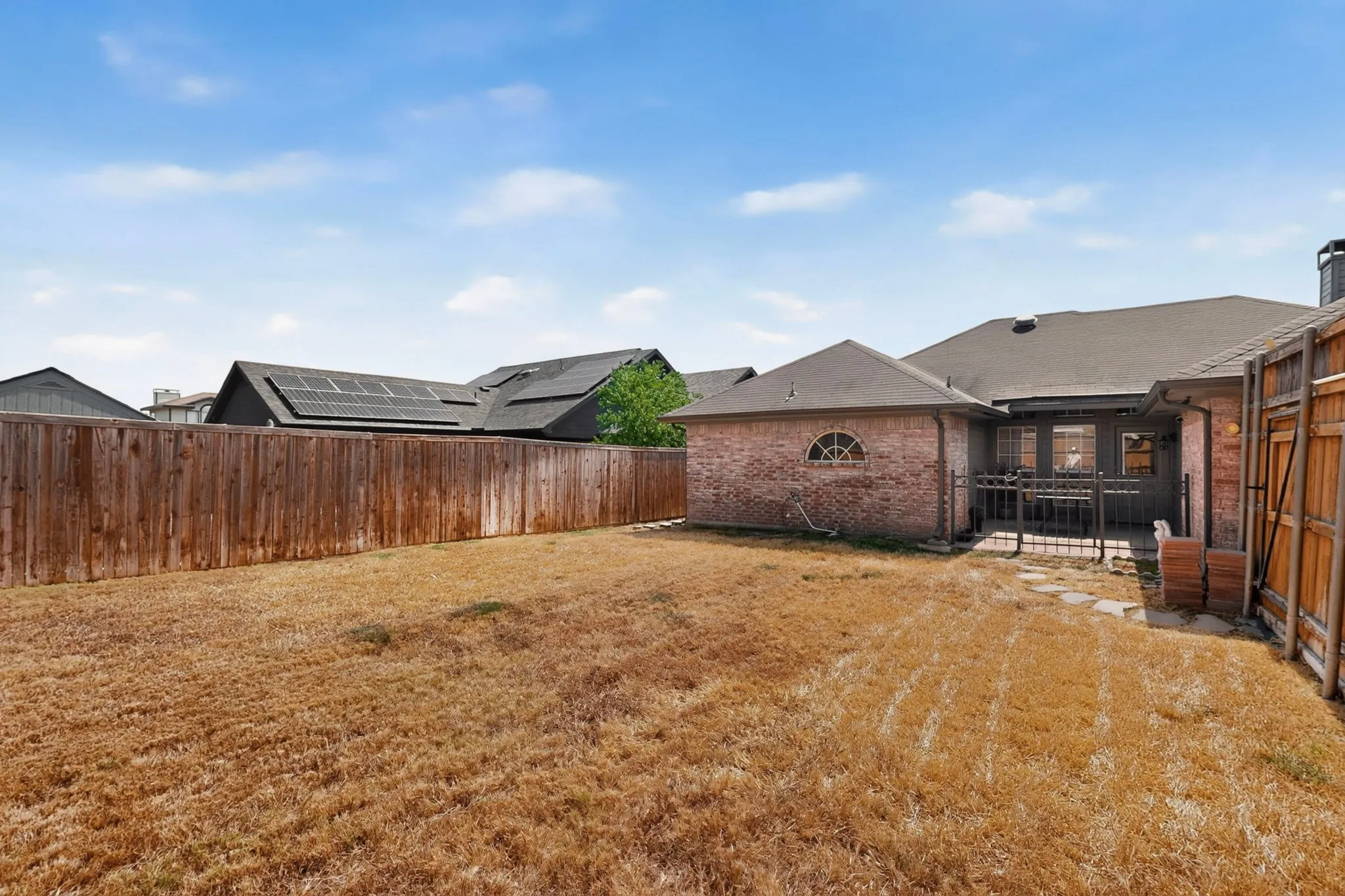 Expansive backyard space featuring a wood privacy fence, a brick exterior structure with an arched window, and a covered patio area with a dark railing