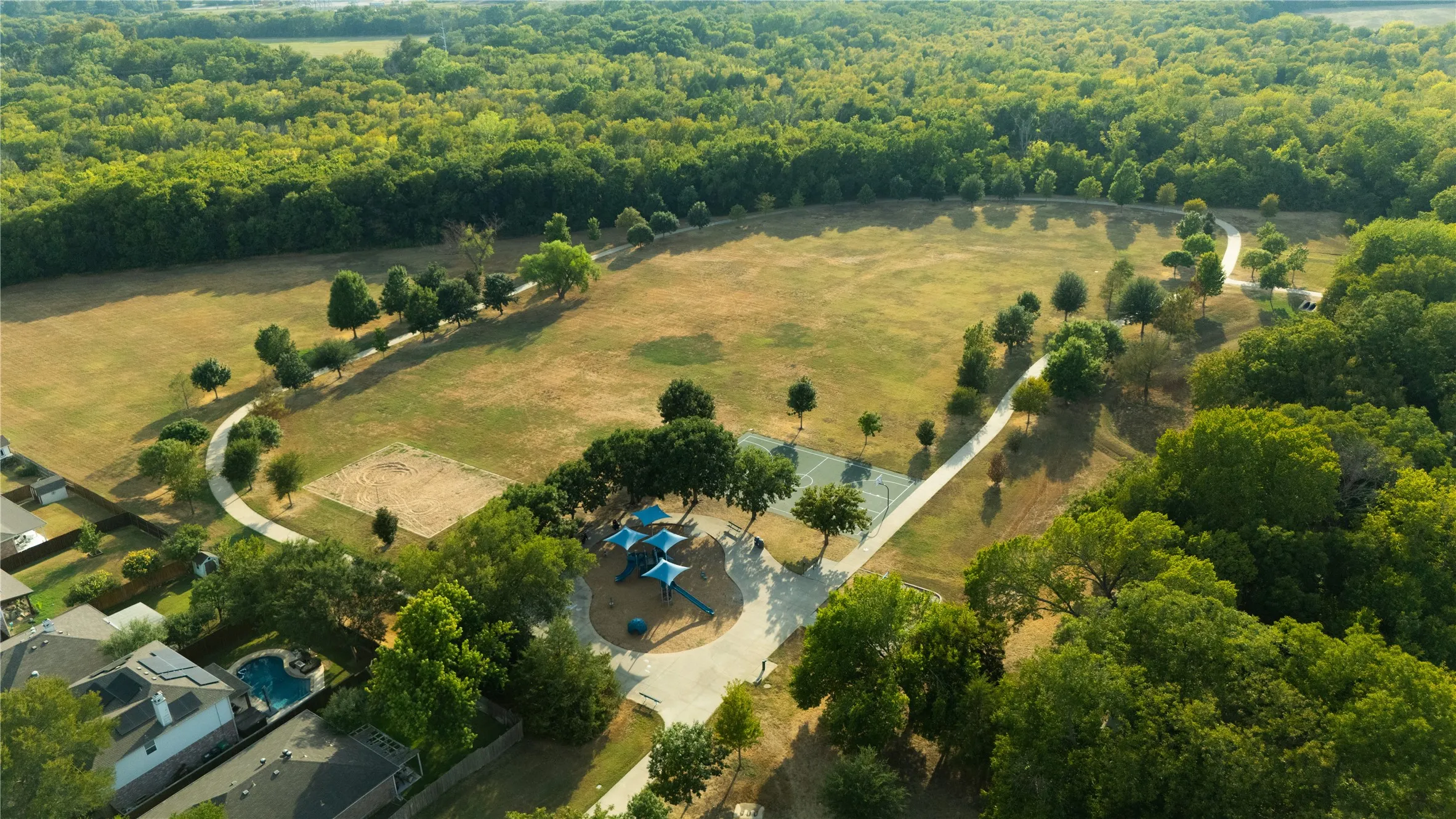 Expansive green space featuring a paved walking path, mature trees, a sand volleyball court, and a recreational playground