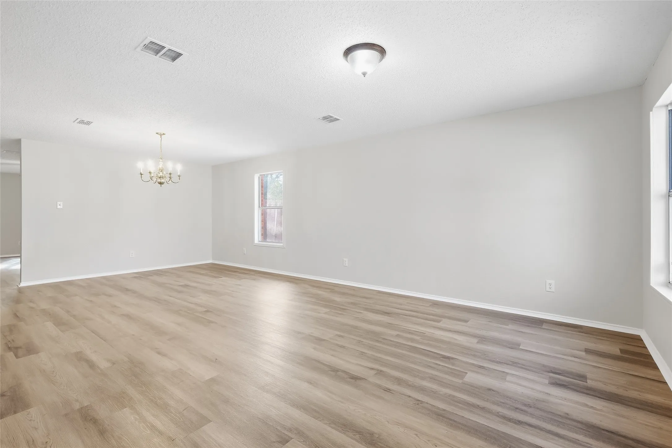Spacious room with wood-finish flooring, two windows, a ceiling-mounted light fixture, and a decorative chandelier