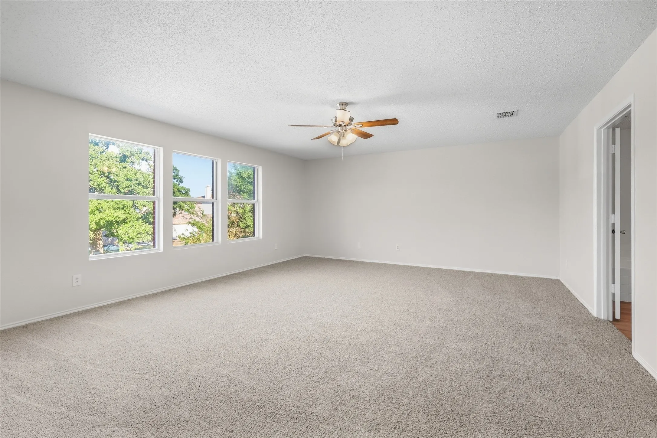 Spacious room featuring light-colored carpet, three windows, a ceiling fan with light, and white walls