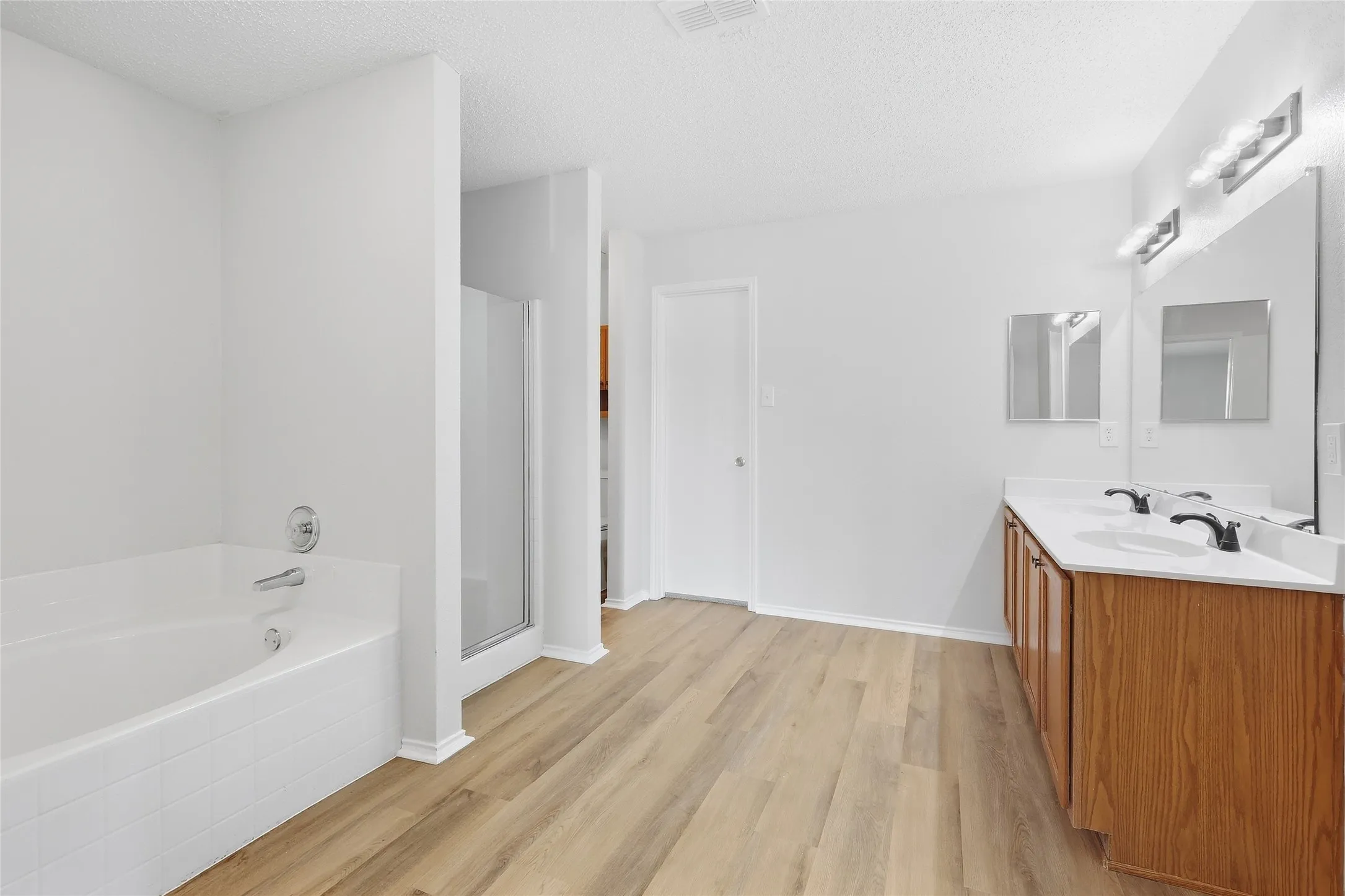 Spacious bathroom featuring a built-in bathtub, glass-enclosed shower, dual vanity with wood-finish cabinetry, and light wood-finish flooring