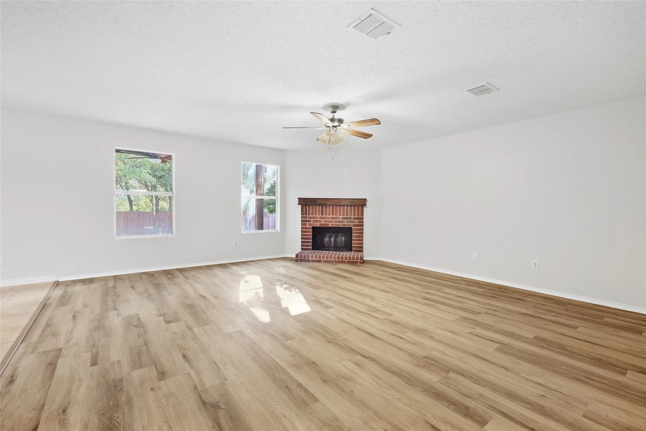 Spacious room featuring wood-finish flooring, white walls, multiple windows, a brick fireplace, and a ceiling fan